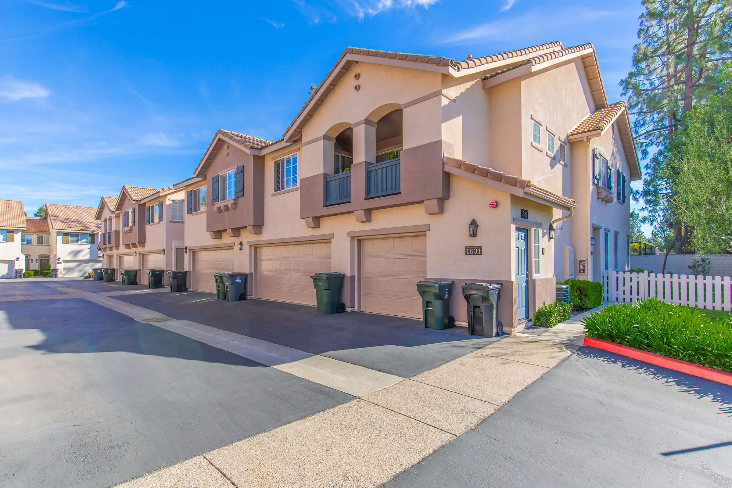 Row of beige townhouses with tile roofs, featuring garages and green waste bins. The scene is set on a sunny day with blue skies and a paved parking area. Lush greenery and a white picket fence are visible in the background. The overall atmosphere is residential and inviting.
