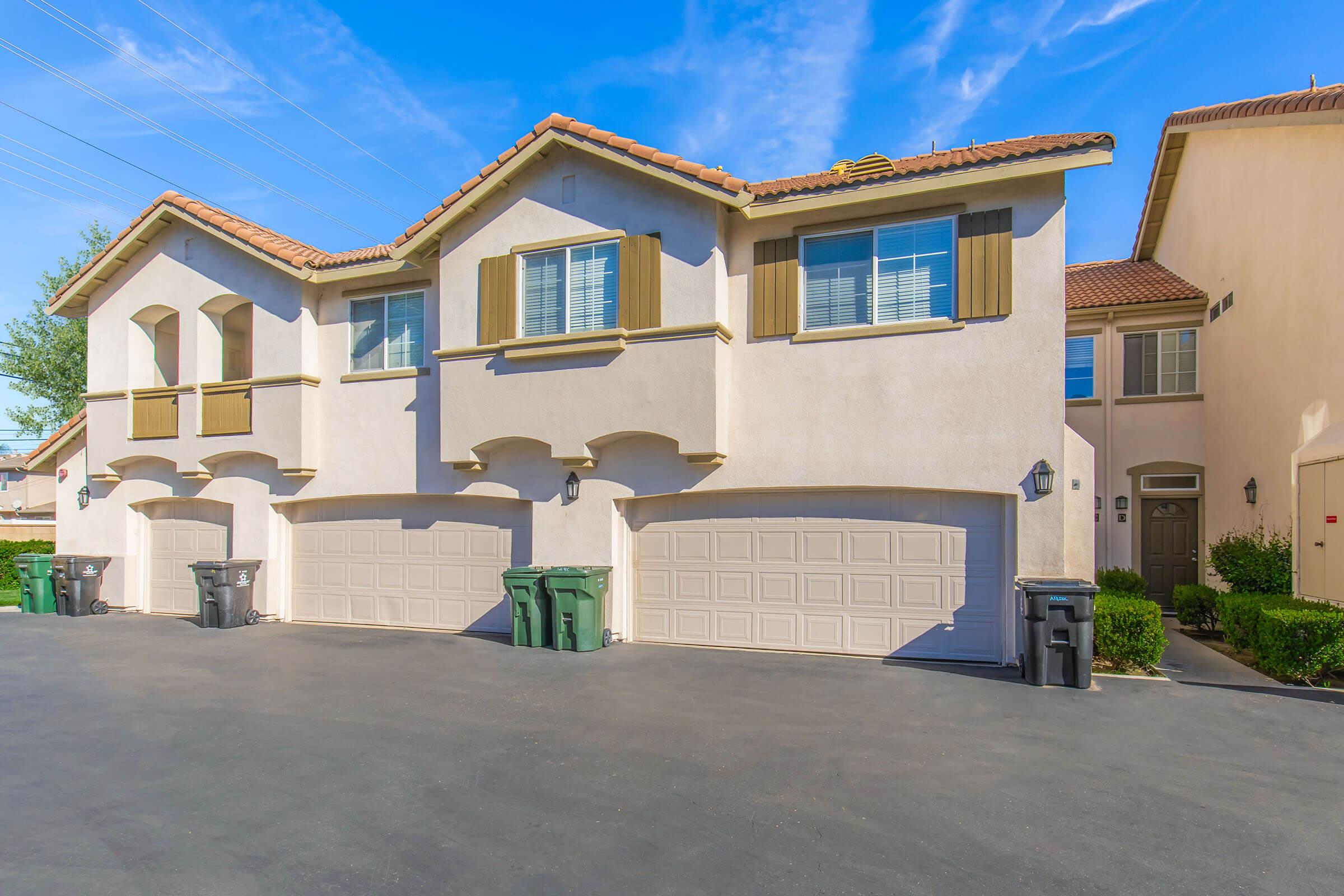 A two-story residential building with beige stucco exterior, featuring windows with shutters and three attached garages. In front of the garages, there are two green trash bins on a paved driveway. The sky is blue with some clouds, and the surrounding area includes greenery.