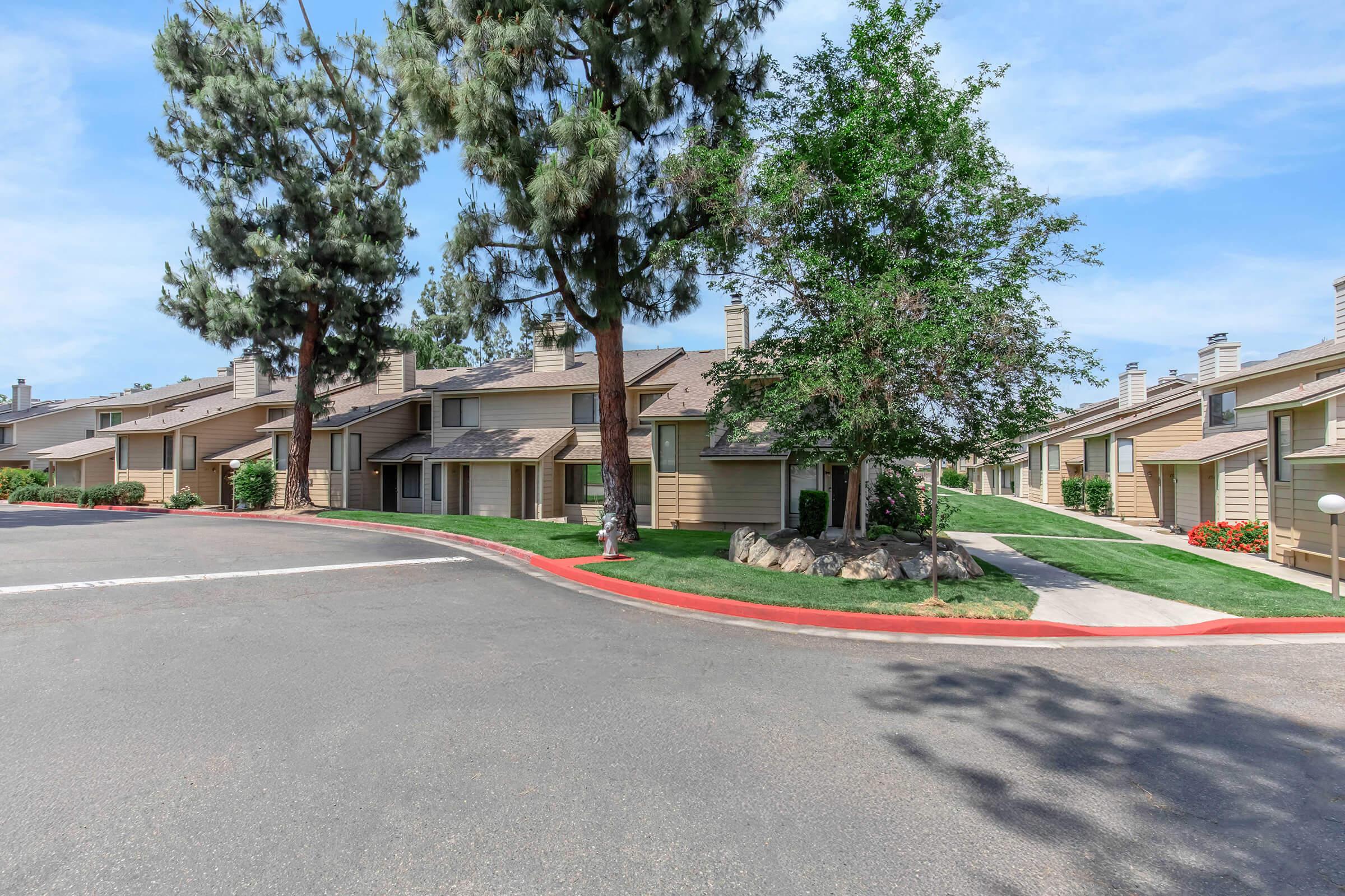 A residential area featuring two-story townhouses surrounded by greenery. There are tall pine trees and well-maintained landscaping with rocks and flower beds along the pathways. The asphalt road curves around the buildings, creating a welcoming atmosphere. Clear blue sky in the background.