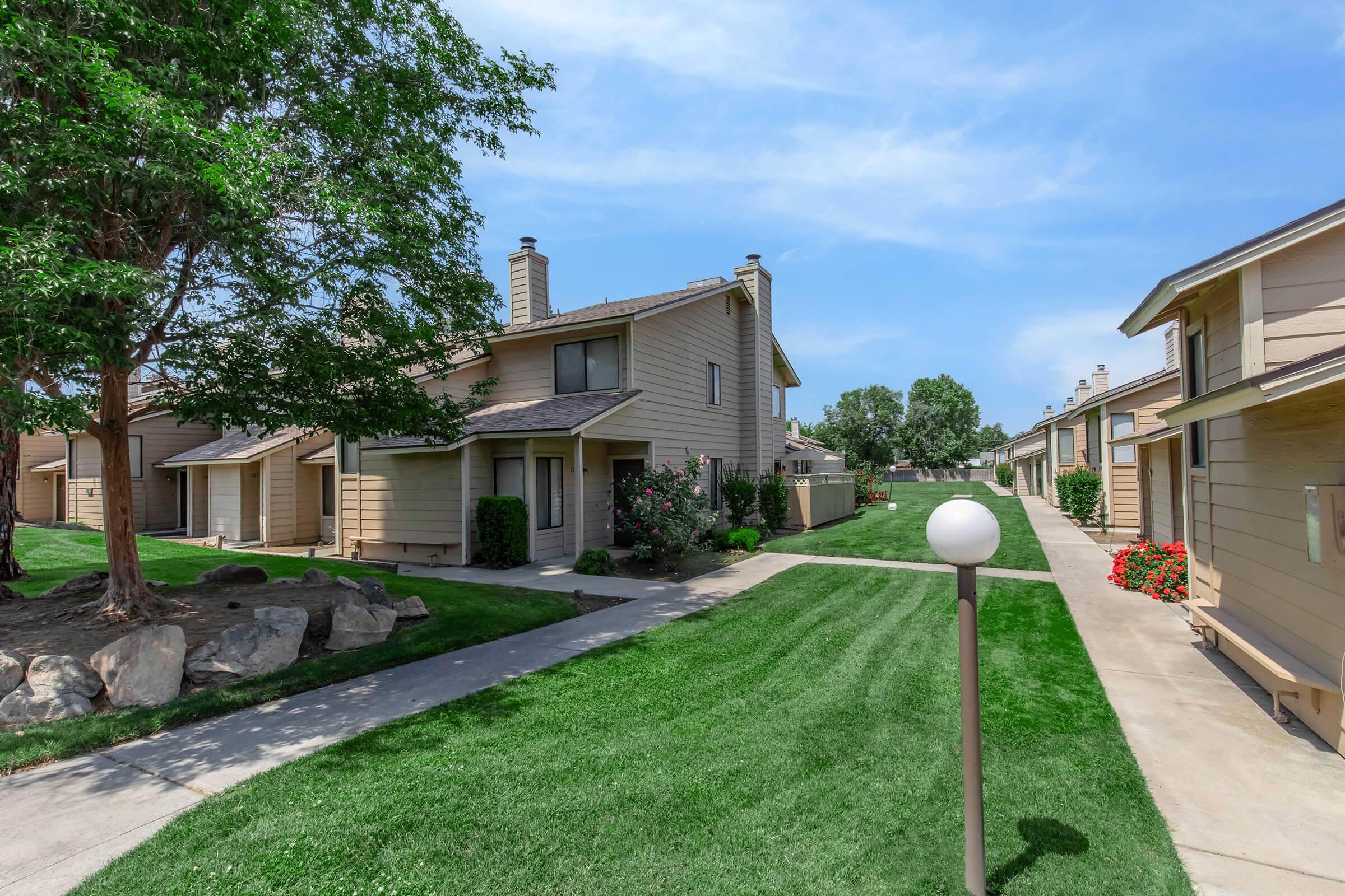 A serene residential area featuring two-story beige apartment buildings surrounded by lush green lawns, trees, and flower beds. A concrete pathway leads through the property, with landscaped gardens and a decorative light post enhancing the inviting atmosphere. Clear blue skies in the background.