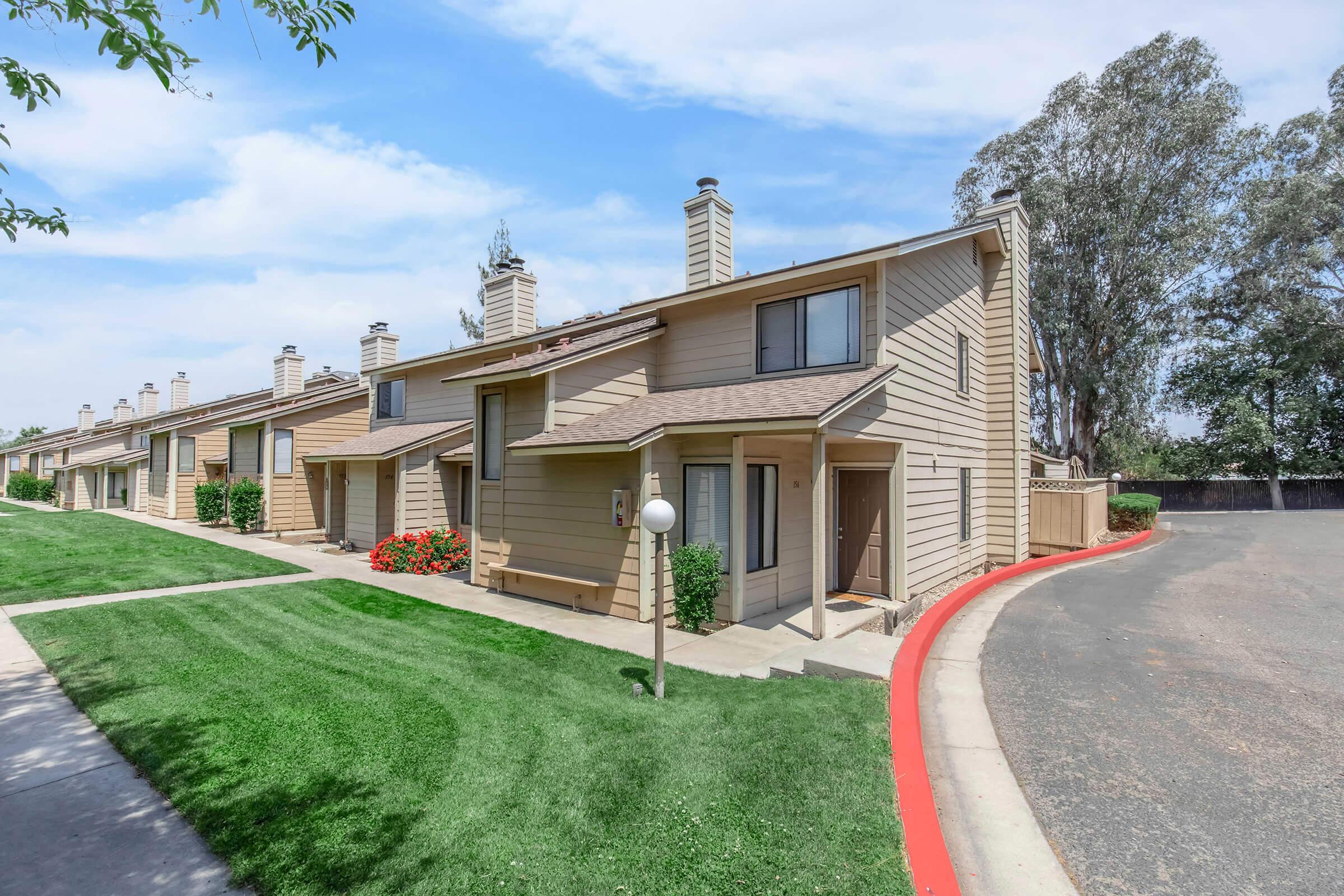 A row of light brown townhouses with pitched roofs and chimney stacks. Each unit features a small front porch and large windows. The neatly landscaped area includes green grass and red flowers, with a curved driveway visible to the right. The sky is partly cloudy, indicating a bright day.