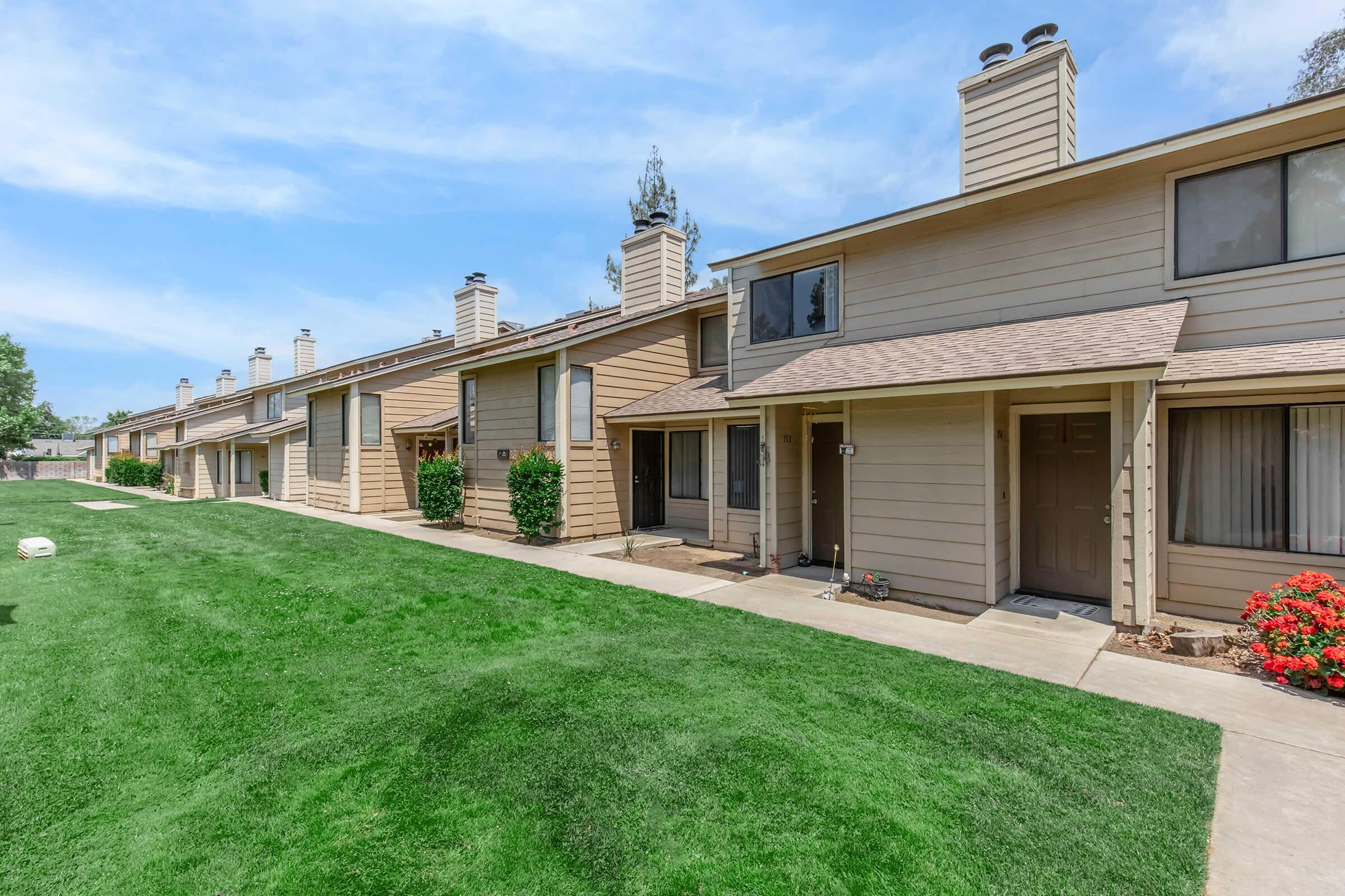 Row of modern two-story townhouses with light brown siding and sloped roofs, set against a green lawn and blue sky. Each unit features a front door with pathway access and small shrubs or flowers near the entrance, creating an inviting suburban atmosphere.