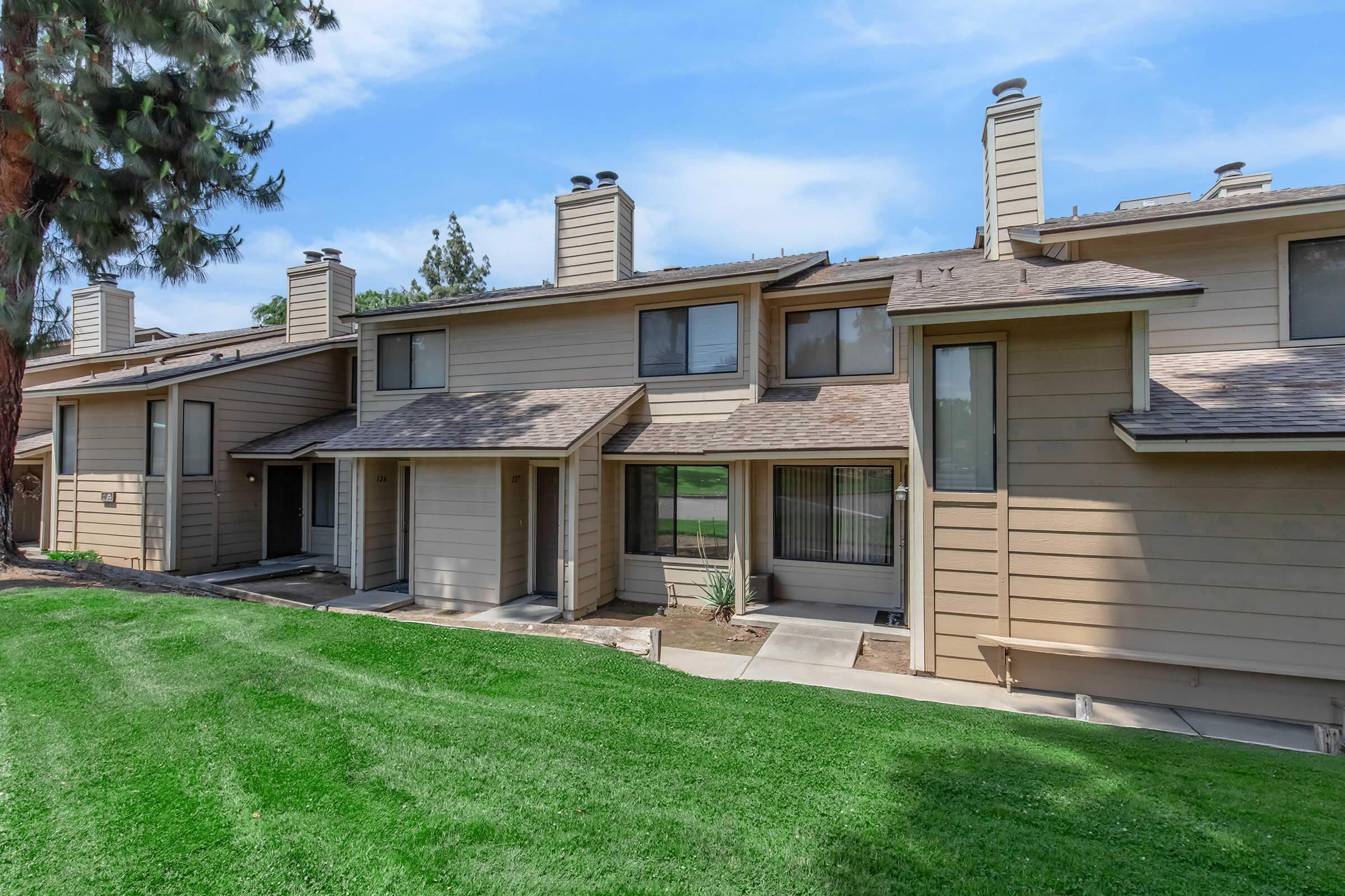 A row of beige townhouse-style buildings with multiple chimneys, set against a green lawn. The architecture features large windows and a simple, modern design. Trees are visible in the background, under a clear blue sky.