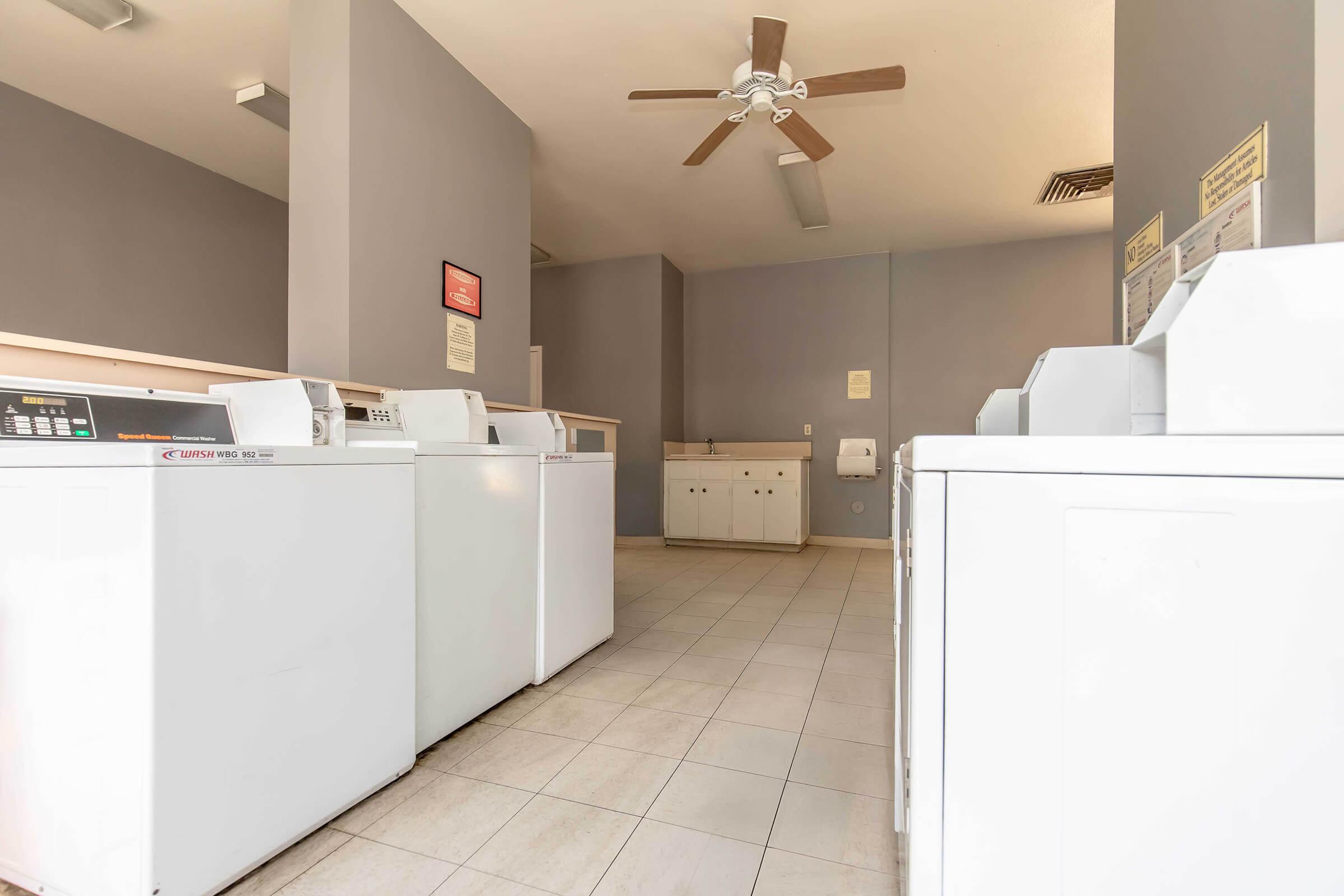A clean and spacious laundromat interior featuring several white washing machines, a ceiling fan, and light gray walls. There is a countertop area with a sink and informational signs on the walls, creating a bright and organized environment for doing laundry.