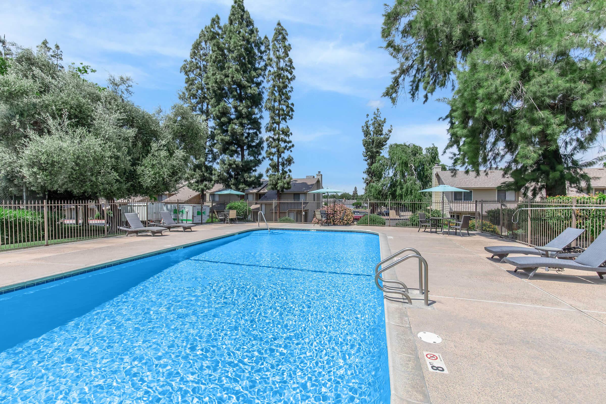 A clear blue swimming pool surrounded by a sunbathing area with lounge chairs. Lush trees and greenery provide a natural backdrop, and nearby there are residential units visible in the distance. The sky is bright and blue, suggesting a sunny day.