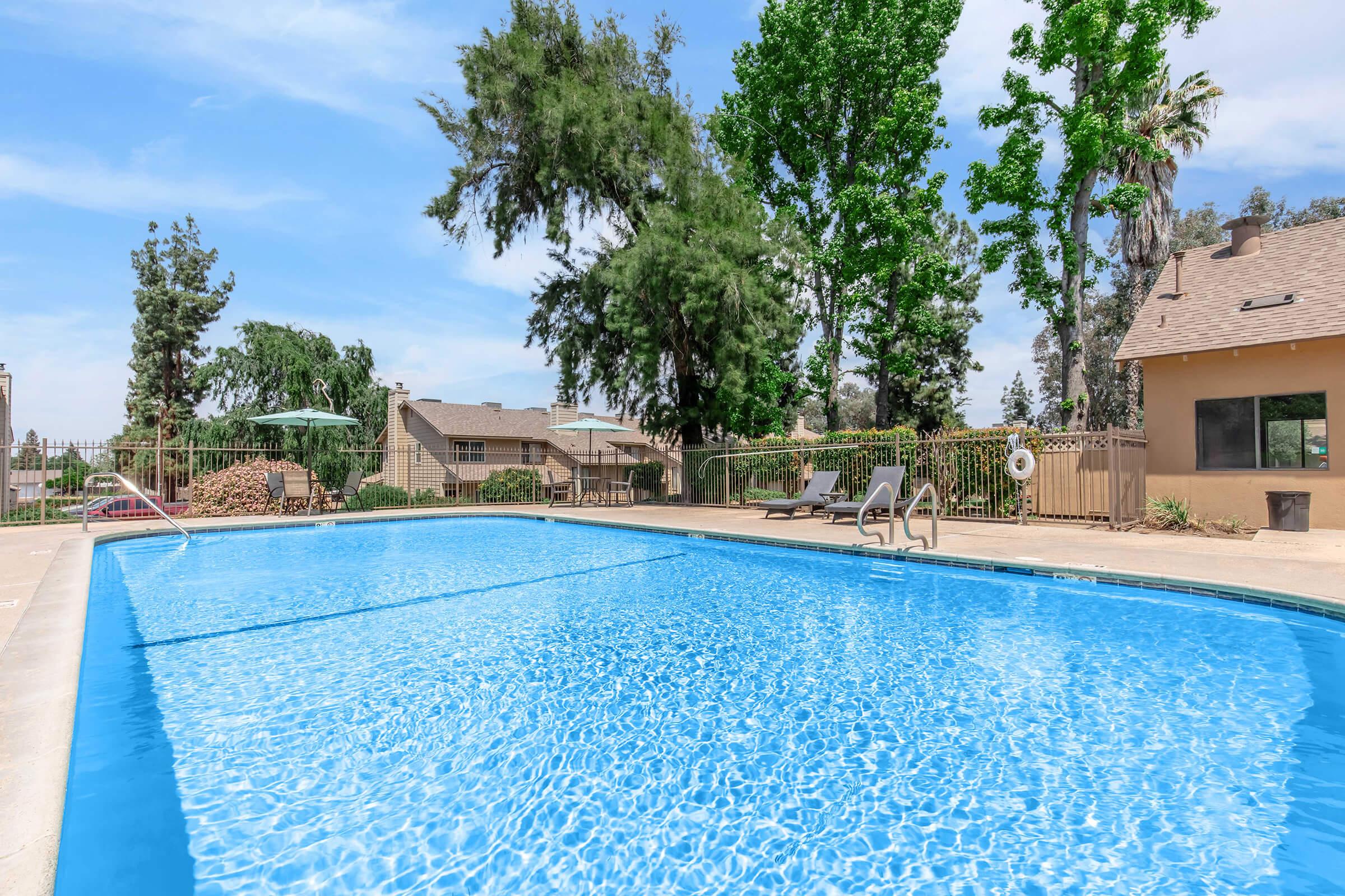 A clear blue swimming pool surrounded by lush green trees and a well-maintained lawn. Sun loungers are positioned near the pool, and there is a shaded umbrella on one side. The backdrop features residential buildings under a bright, sunny sky.