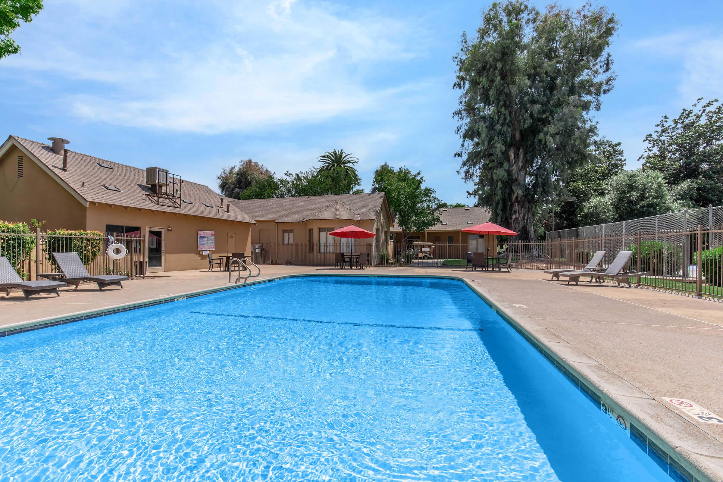 A clean swimming pool surrounded by lounge chairs, with a few red umbrellas providing shade. In the background, there are trees and a building with outdoor amenities visible, under a clear blue sky. The setting is inviting and designed for relaxation and recreation.