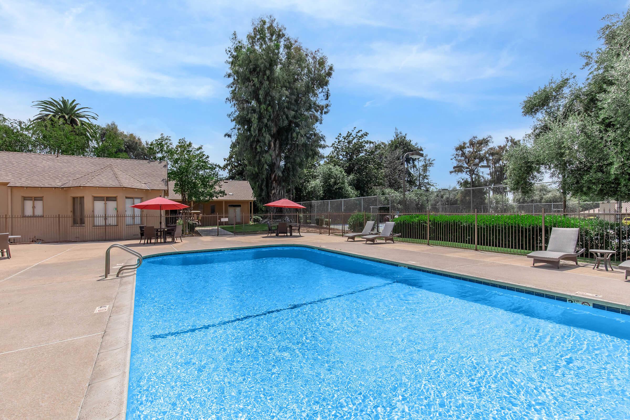 A clear blue swimming pool surrounded by a landscaped area with lounge chairs and red umbrellas. In the background, there are trees and a building, contributing to a relaxing outdoor environment. The scene is bright and inviting, perfect for leisure and enjoyment.