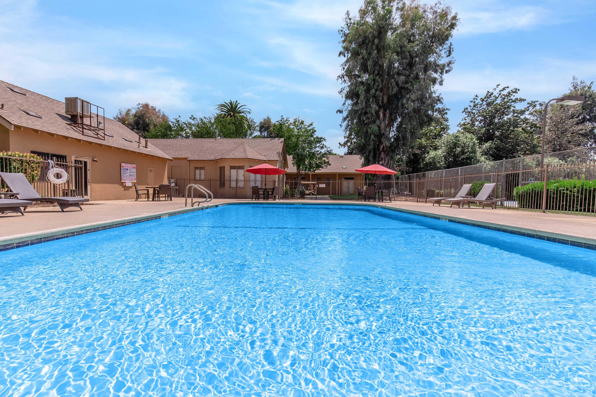 A sparkling blue swimming pool surrounded by lounge chairs and red umbrellas. In the background, a beige building with trees and landscaped greenery can be seen under a clear blue sky. The atmosphere is tranquil and inviting, perfect for relaxation.