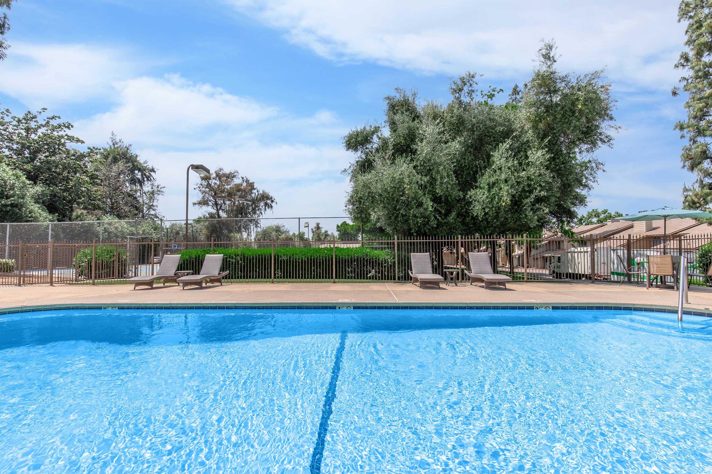 A sparkling blue swimming pool with lounge chairs nearby, surrounded by a fenced area. Lush greenery and trees can be seen in the background under a clear blue sky, creating a relaxing outdoor space suitable for leisure and recreation.