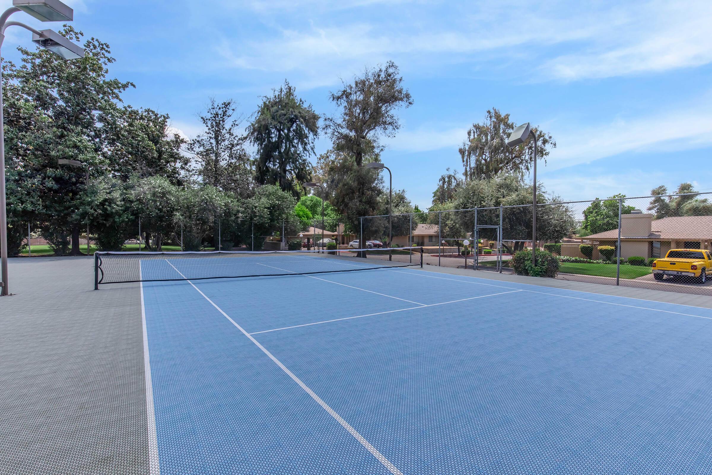 A view of a well-maintained outdoor tennis court with blue surfaces, surrounded by trees and residential buildings. The court is empty, with clear blue skies and bright lighting, indicating a sunny day. A yellow vehicle is parked nearby.