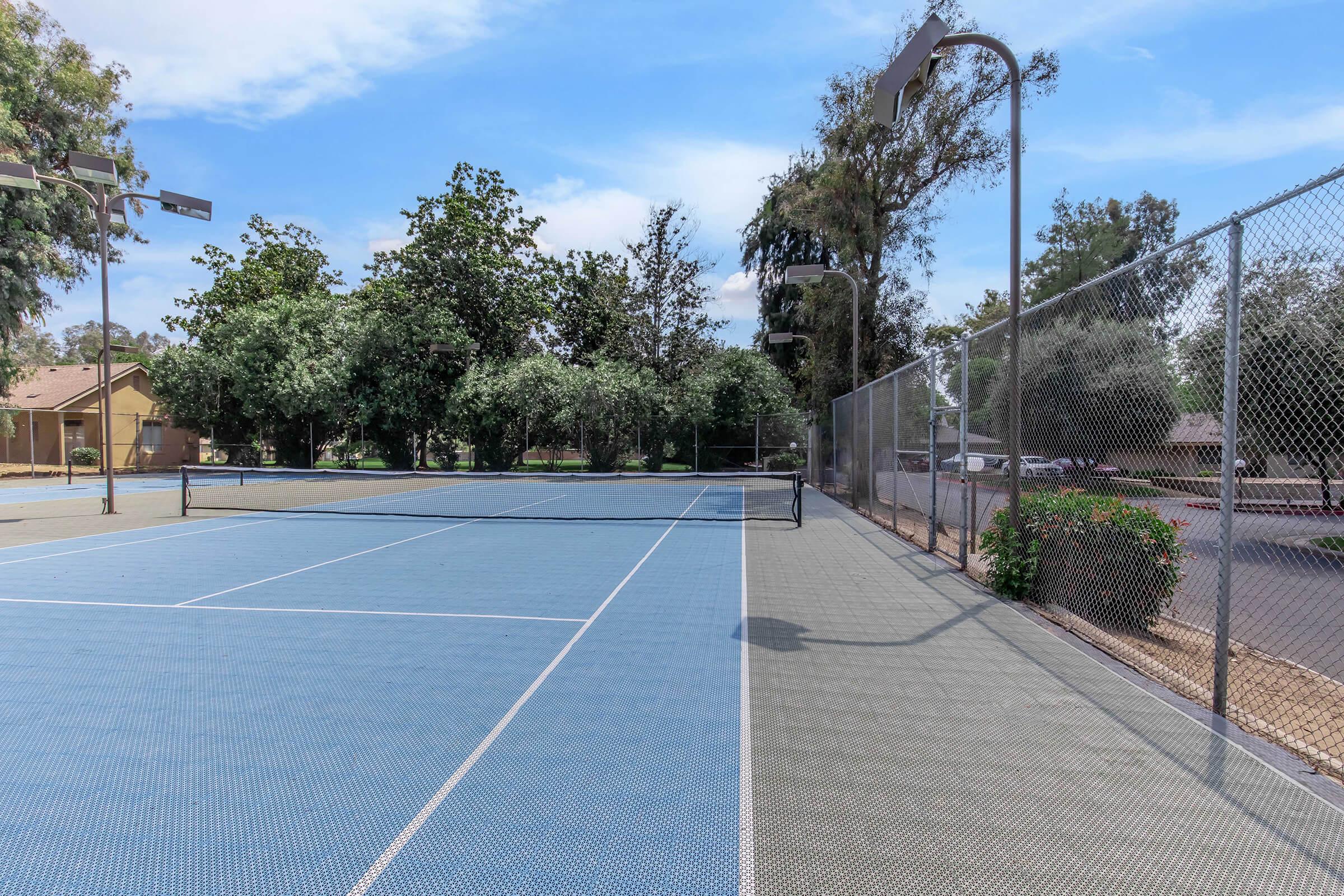 A tennis court featuring a blue playing surface, surrounded by trees and greenery. There is a fence along one side and outdoor lighting fixtures. Nearby parking is visible in the background, showcasing a peaceful recreational environment.