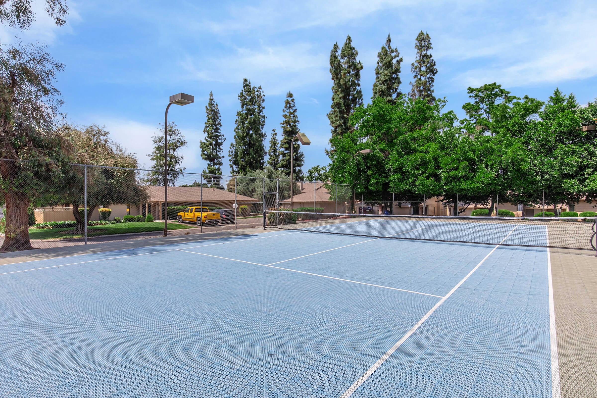 A well-maintained outdoor tennis court with blue surface, surrounded by green trees and shrubs. In the background, there are residential buildings and a yellow vehicle parked nearby. Bright, clear sky above with a few clouds.