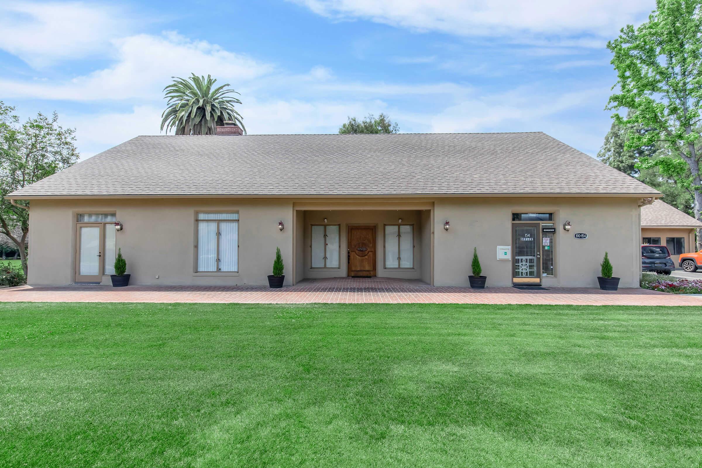 A single-story house with a light beige facade, a central front door with a decorative wood design, and large windows on either side. The front yard features well-manicured green grass and two small conical shrubs. Palm trees are visible in the background against a clear blue sky.