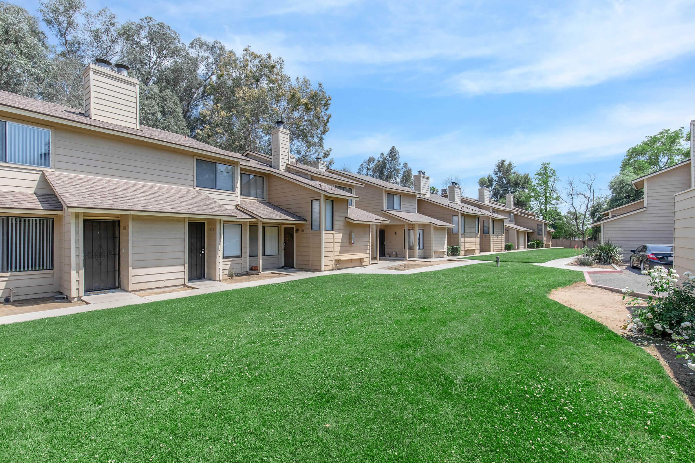Row of beige townhouses with brown roofs, arranged along a grassy pathway. Each unit has a small porch and windows with vertical blinds. Lush greenery surrounds the area, with a few trees in the background under a clear blue sky.