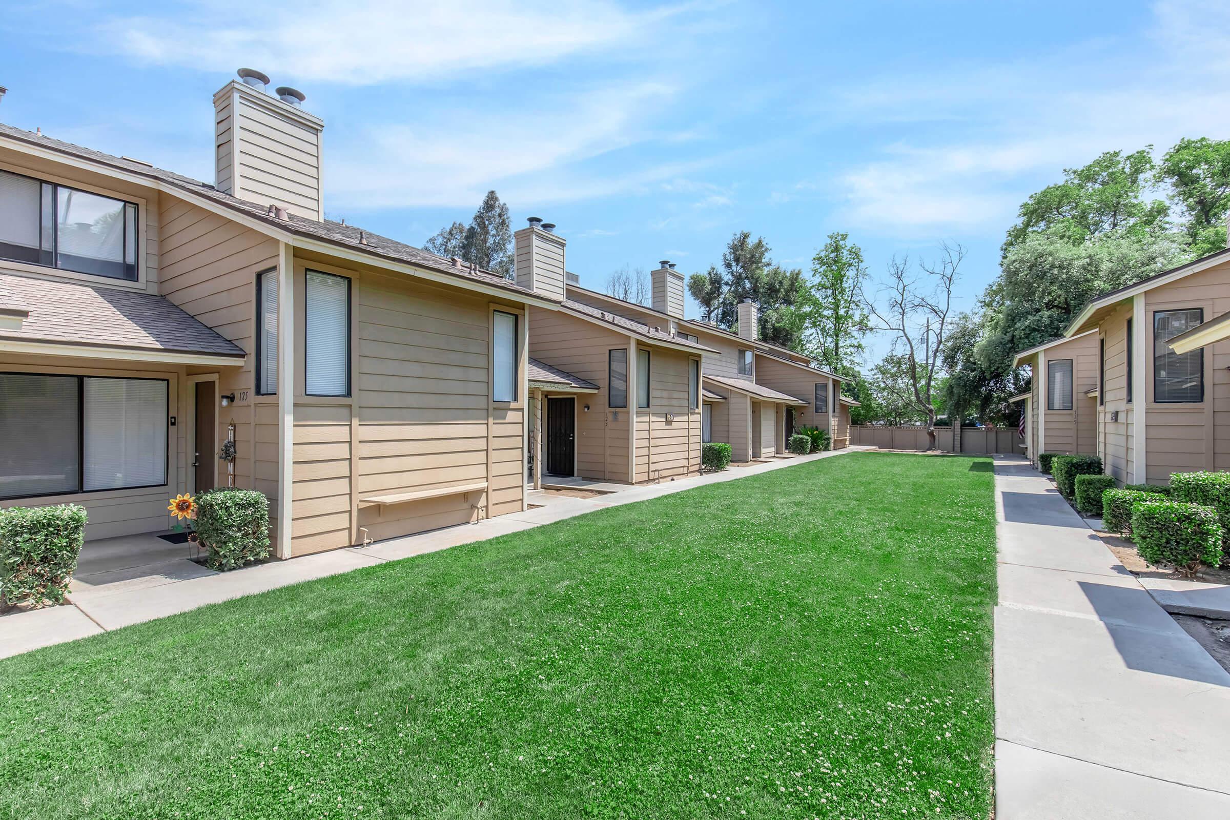 A sunny day view of a residential area featuring two rows of beige apartment buildings. Each unit has a front porch and chimneys, with well-maintained green lawns and hedges in between the pathways. Trees and clear blue skies add to the peaceful ambiance.
