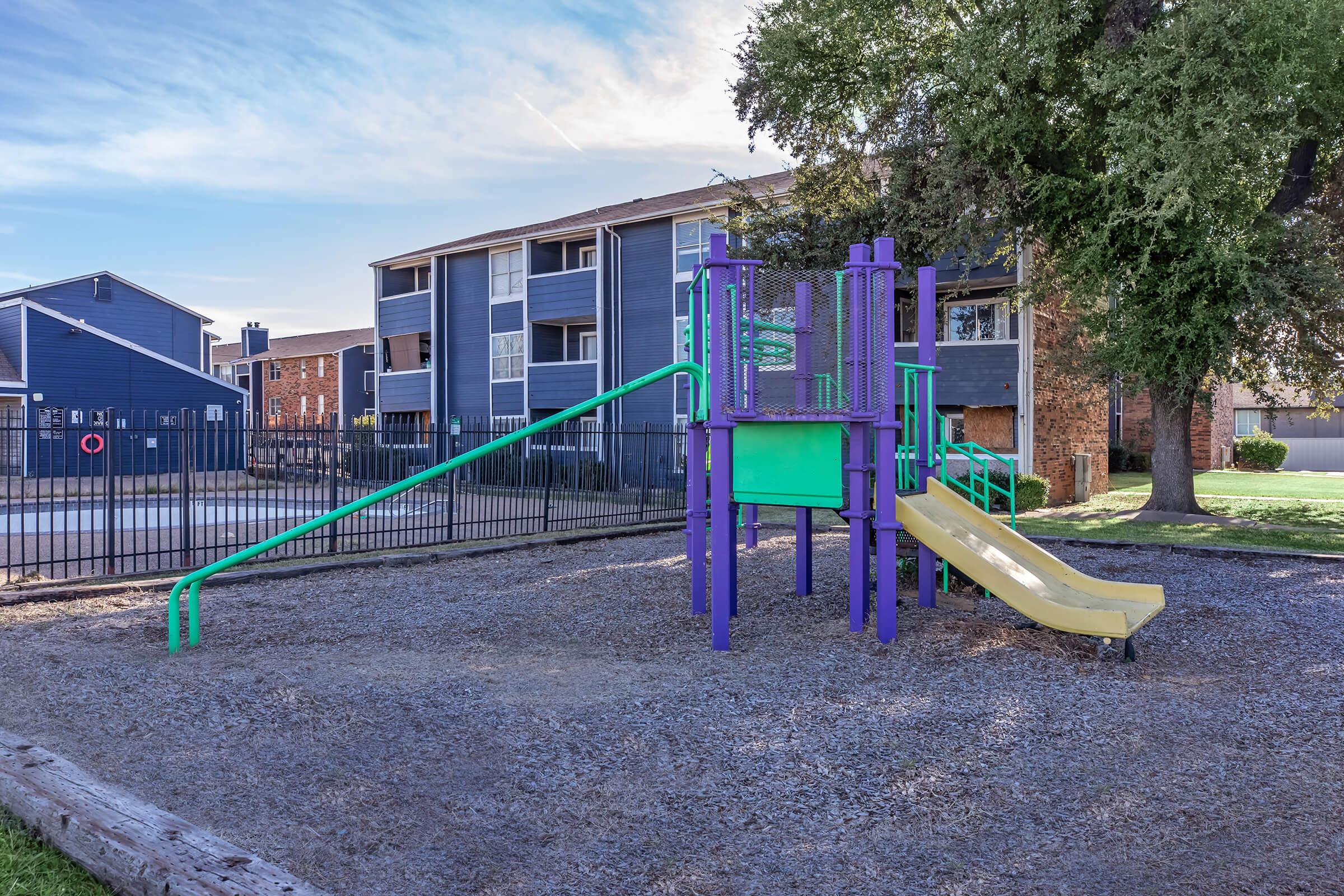 A colorful playground structure featuring a slide and climbing elements, surrounded by gravel. In the background, there are residential buildings with blue façades and a fenced area, with trees providing shade nearby.