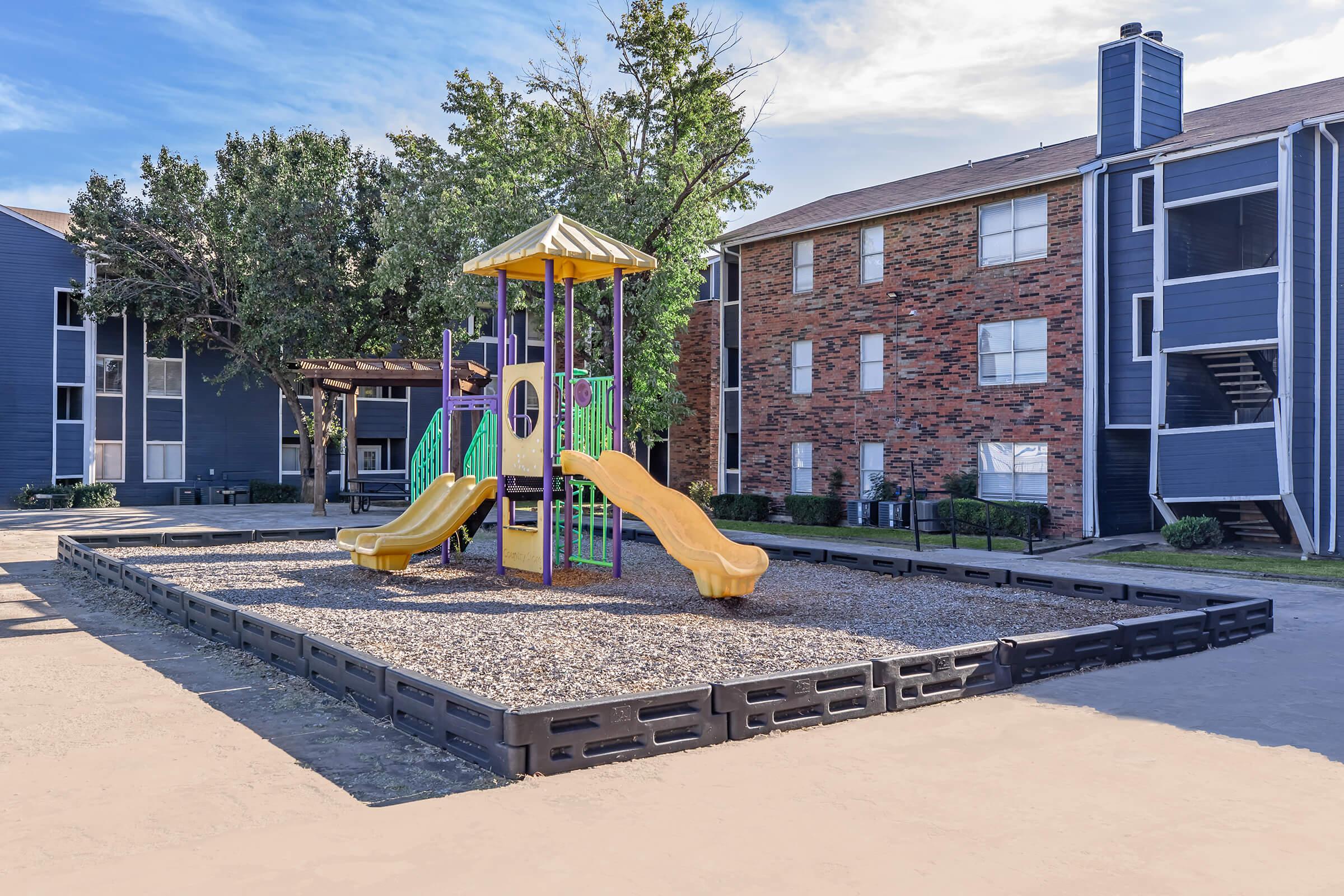 Playground area featuring two yellow slides and a colorful climbing structure, surrounded by gravel. In the background, there are two apartment buildings with a mix of brick and blue exteriors, under a clear sky with a few trees providing shade.