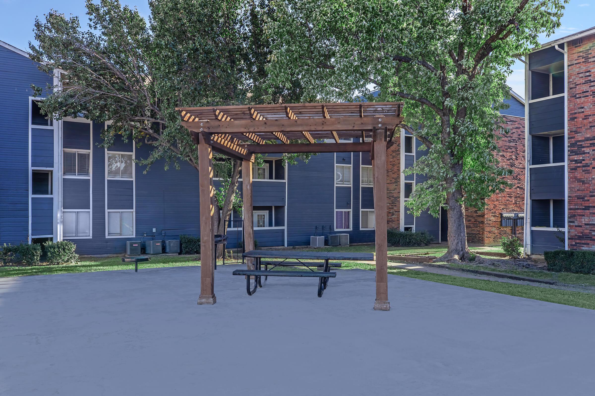 A wooden pergola with a picnic table underneath, situated in a courtyard surrounded by apartment buildings. Lush green trees are visible, contributing to the outdoor setting. The ground is paved, and the area appears well-maintained and inviting for relaxation or gatherings.