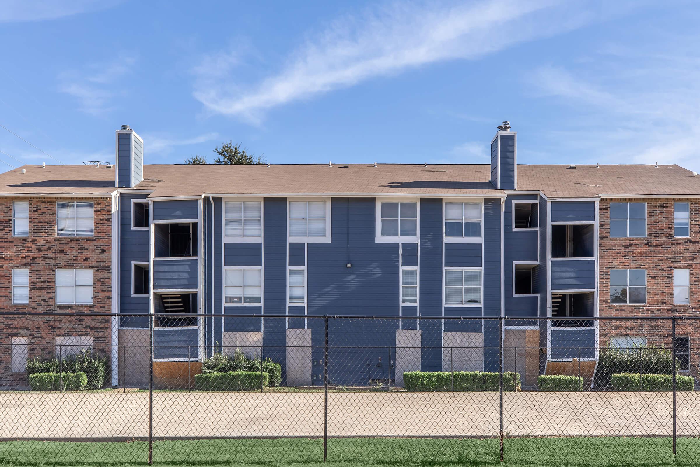 A blue and brick apartment building with multiple balconies, surrounded by a green lawn and a chain-link fence. The sky is clear with some wispy clouds. The building features large windows and a symmetrical design.