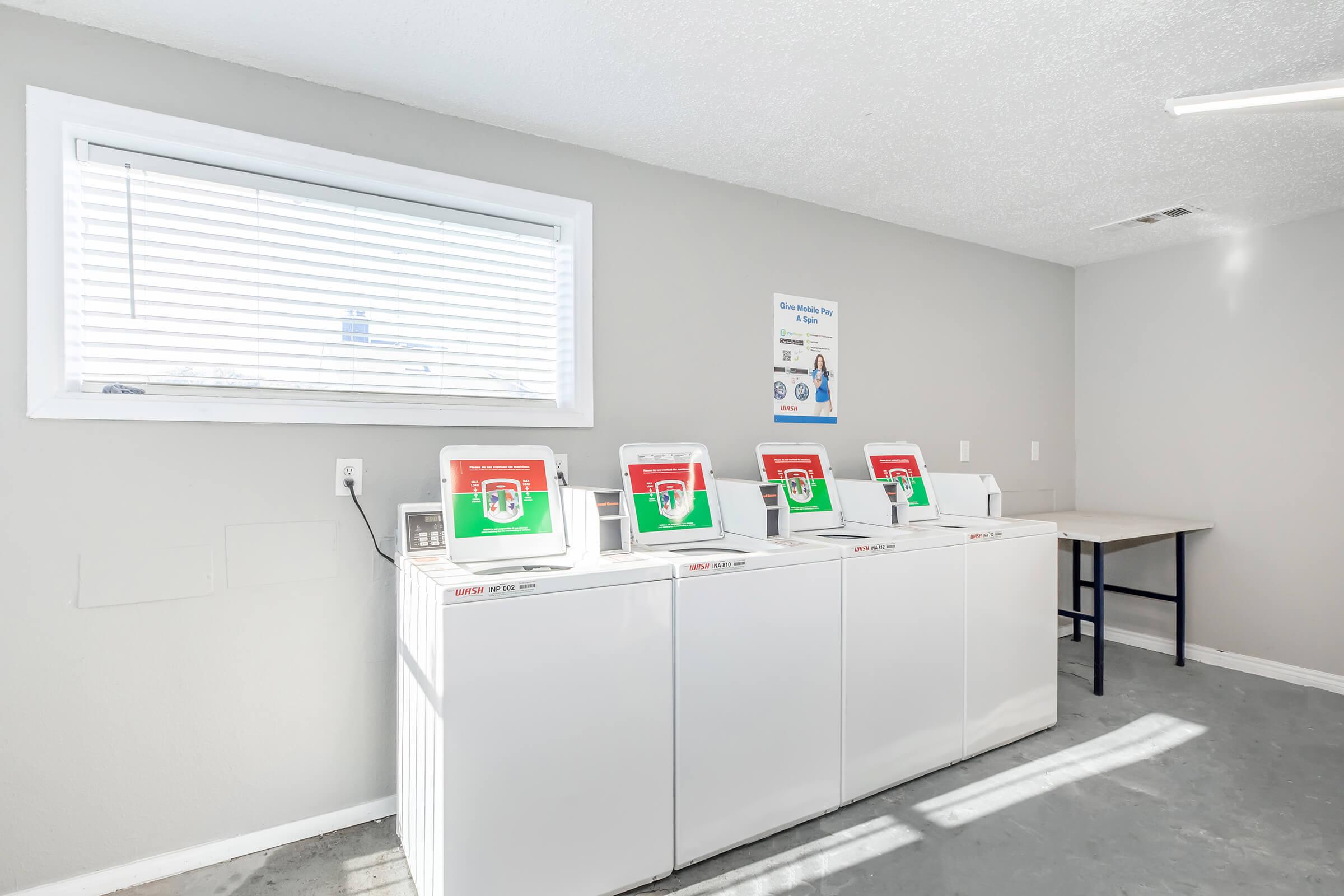 A clean and well-lit laundry room featuring four white washing machines with digital displays and green indicators. A large window lets in natural light, and there is a table against the wall. A sign is visible in the background, providing instructions or information about the laundry facilities.