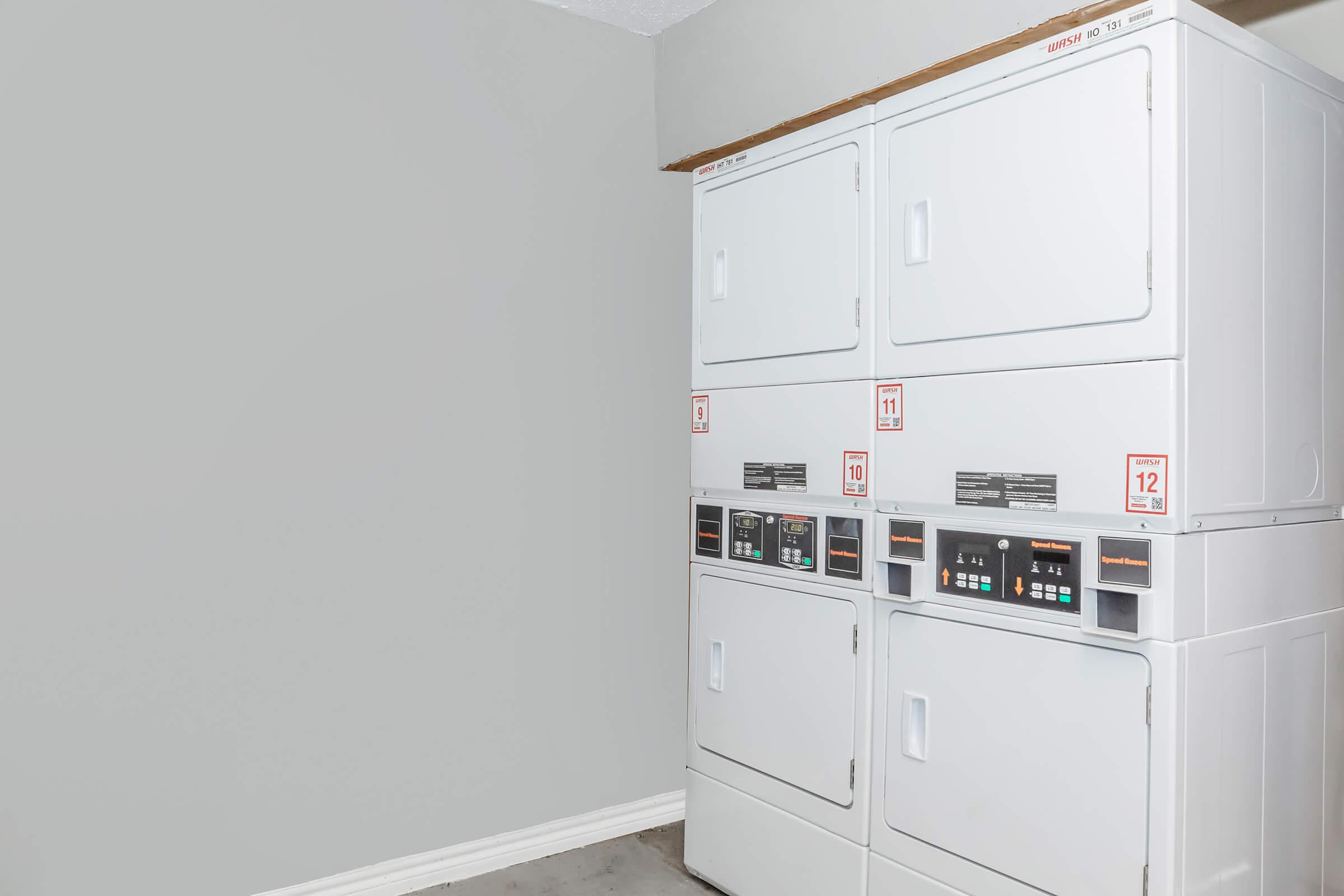 A stacked washer and dryer unit against a gray wall, featuring control panels with buttons and digital displays. The laundry appliances are white and positioned in a corner of a room with a clean and minimalistic appearance.