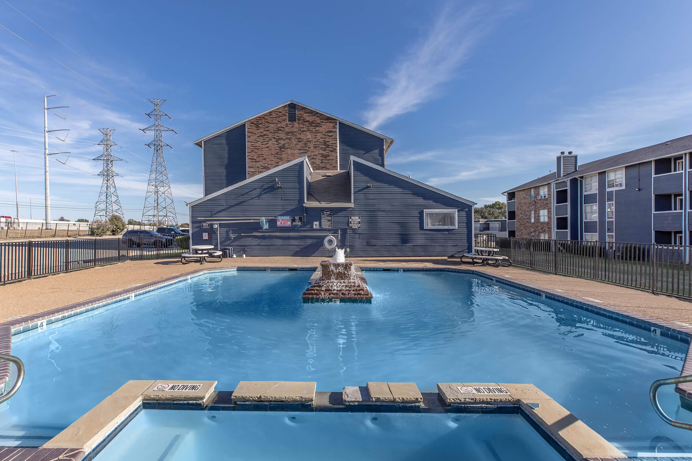 Outdoor swimming pool area with a fountain in the center, surrounded by a fence. In the background, a multi-story building with a modern design and power lines visible against a clear blue sky. Sunlight reflects on the water, creating a bright and inviting atmosphere.