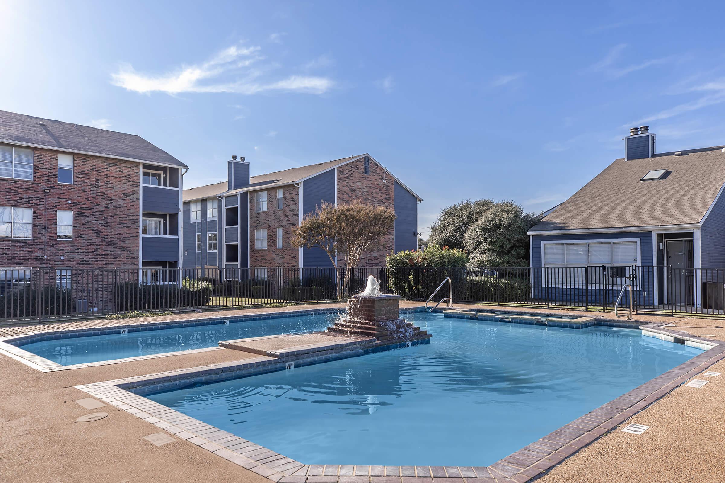 A view of a swimming pool surrounded by residential buildings. The pool features a stone fountain in the center and has a fenced perimeter. Sunlight reflects off the water, and the area is landscaped with trees and grass. The setting conveys a peaceful atmosphere perfect for relaxation.