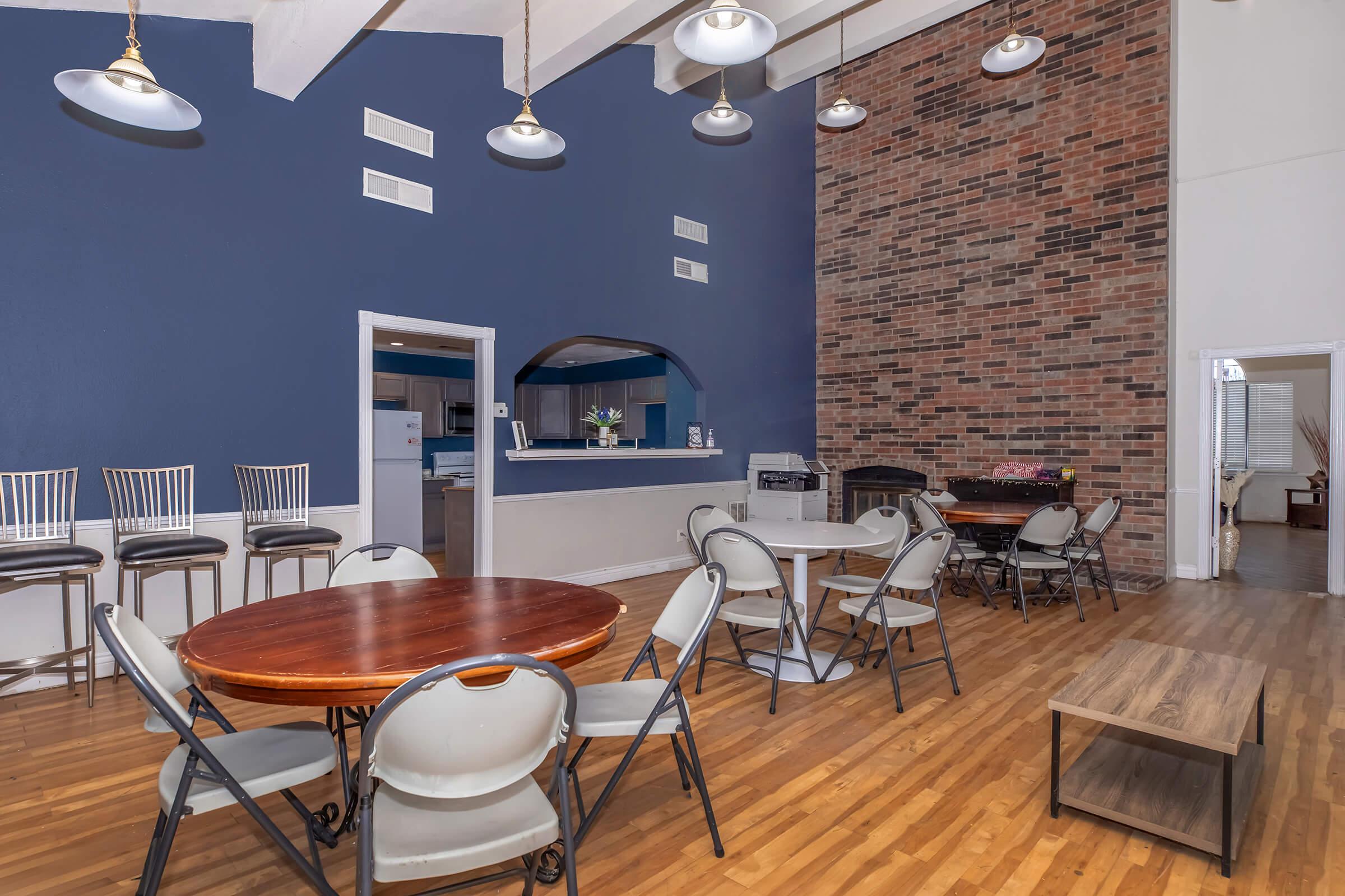 Interior of a spacious room featuring a mix of round and rectangular tables with chairs, a bar area with stools, and a brick accent wall. The space has wooden flooring and well-lit with pendant lights, creating a welcoming atmosphere suitable for gatherings or events.