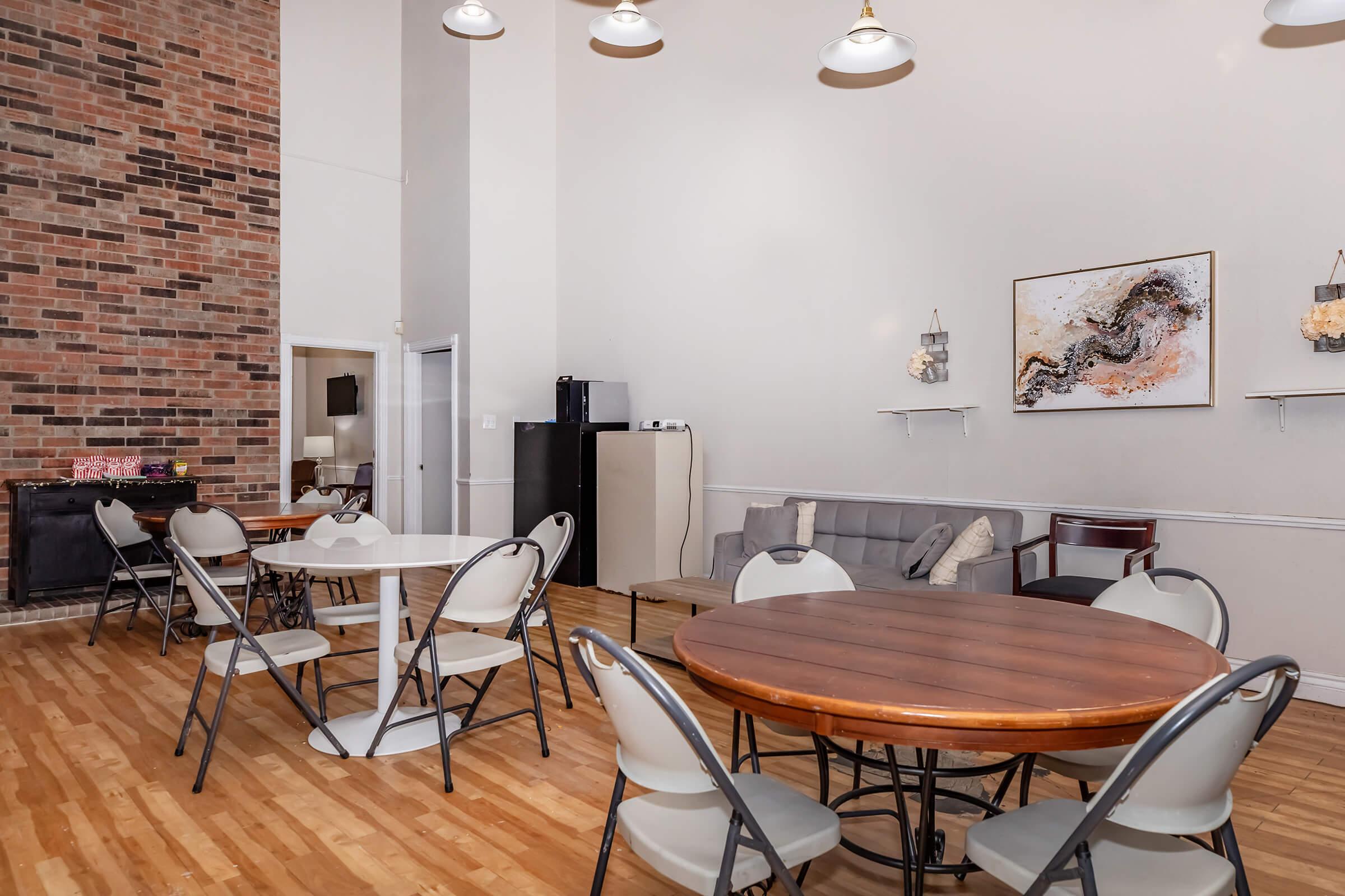 Spacious room with high ceilings featuring several round wooden and white tables surrounded by folding chairs. A brick accent wall adds warmth. In the background, there is a gray sofa, a small kitchenette with a refrigerator, and wall decorations, including a modern art piece.