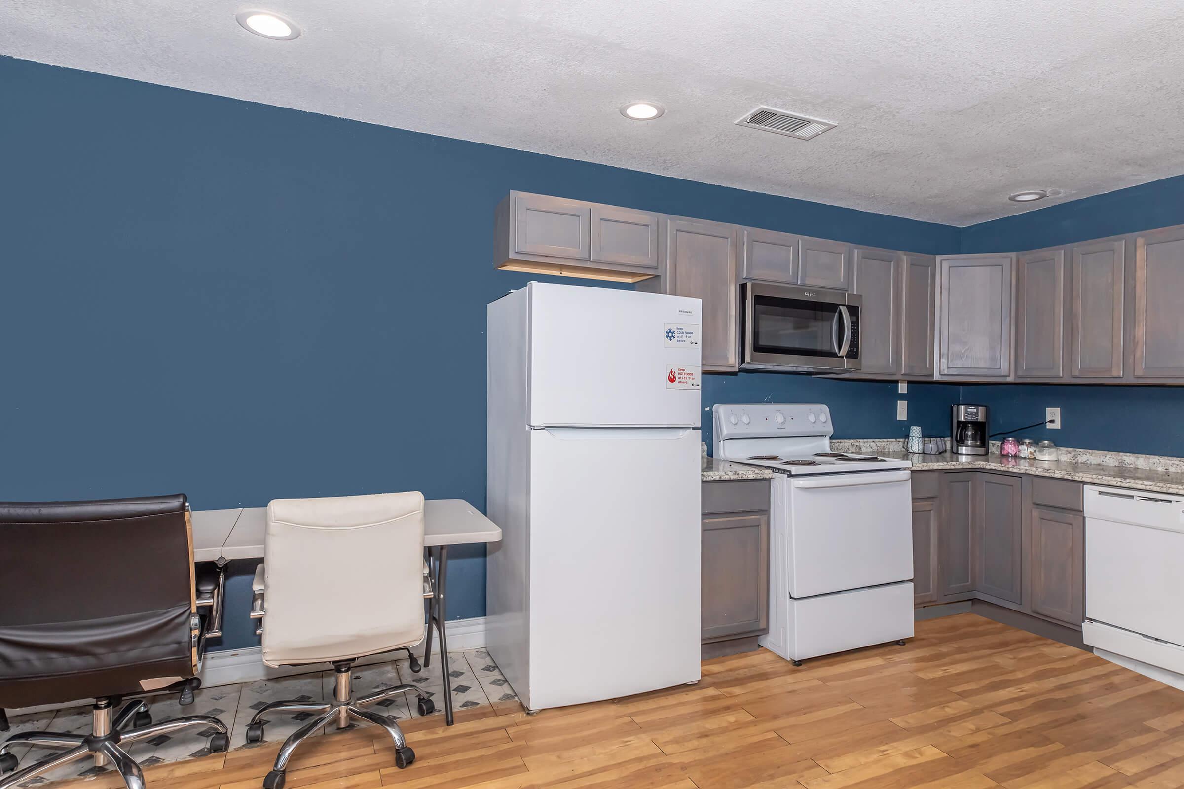 A modern kitchen with light gray cabinets, a white refrigerator, and an electric stove. There's a microwave mounted above the stove and a coffee maker on the counter. A small dining area features a table with two chairs. The walls are painted blue, and the flooring is wooden.