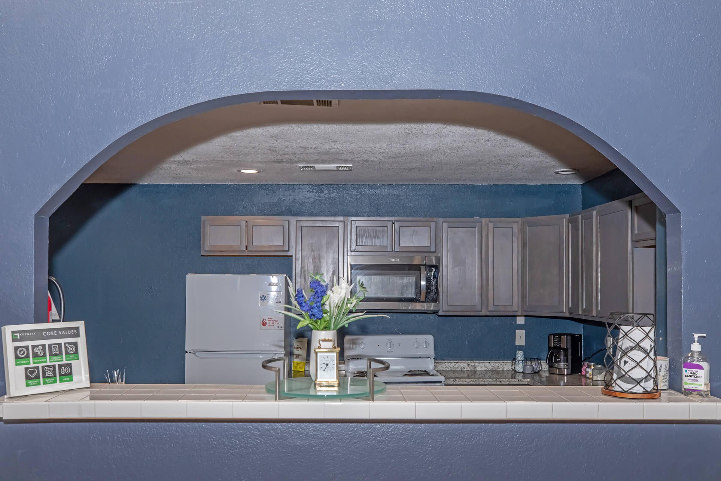 A view of a kitchen through an archway, featuring dark cabinets and a light countertop. A vase with blue flowers sits on the counter, alongside a sign listing community features and a bottle of hand sanitizer. The back wall is painted blue, creating a cozy atmosphere.