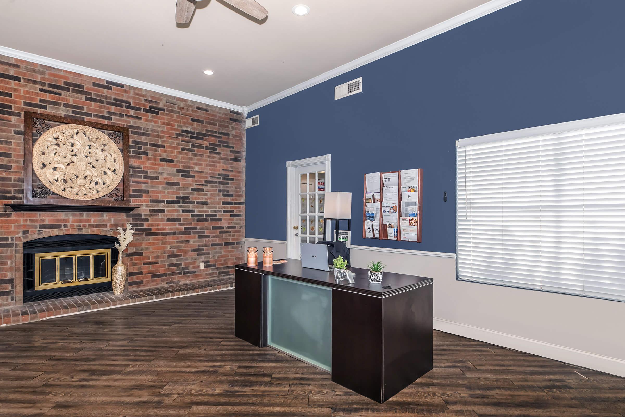 A modern office space featuring a dark wooden desk with a lamp and small plant, a brick accent wall, a fireplace, and a large circular wall decoration. Natural light streams in through a window with white blinds, and there are bulletin boards on the wall.
