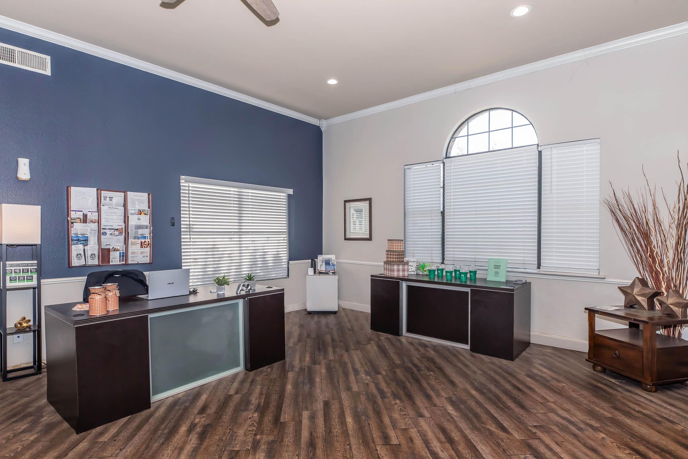 A modern office reception area featuring a dark wood desk with a computer, a water cooler, and informational boards on the wall. Large windows with closed blinds provide natural light. Decorative elements include a tall vase and a coffee station on the side, with hardwood flooring adding warmth to the space.