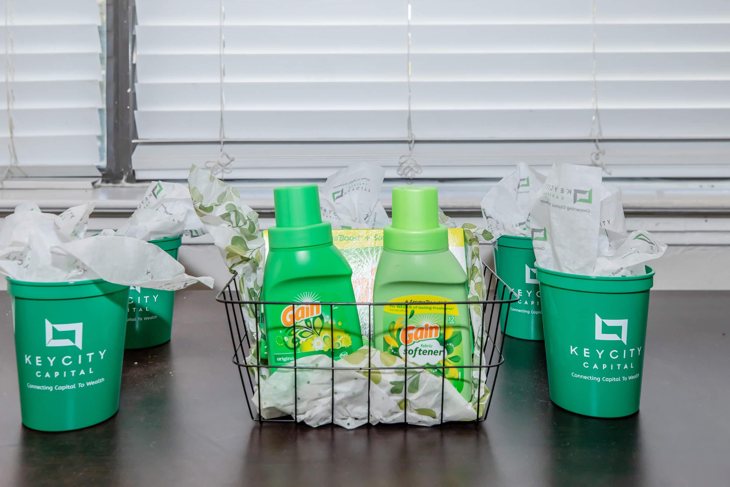 A wire basket containing two bottles of laundry detergent and fabric softener, surrounded by green cups labeled "KEYCITY CAPITAL." The cups are filled with crumpled tissue paper, and the background features a window with blinds.