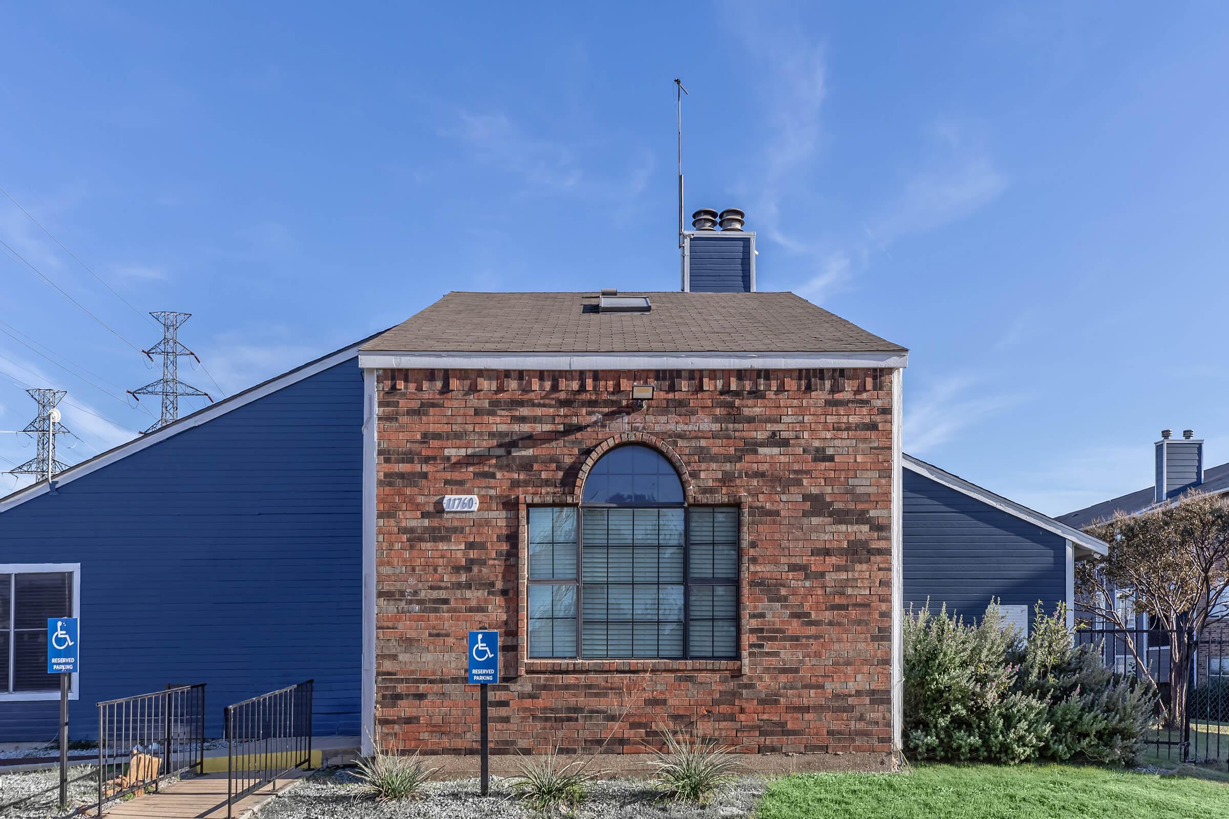 A brick building with a blue panel extension, featuring a large arched window and a chimney on the roof. In front, there is a pathway leading to a ramp with two accessible parking signs. The sky is clear and blue, and power lines are visible in the background.