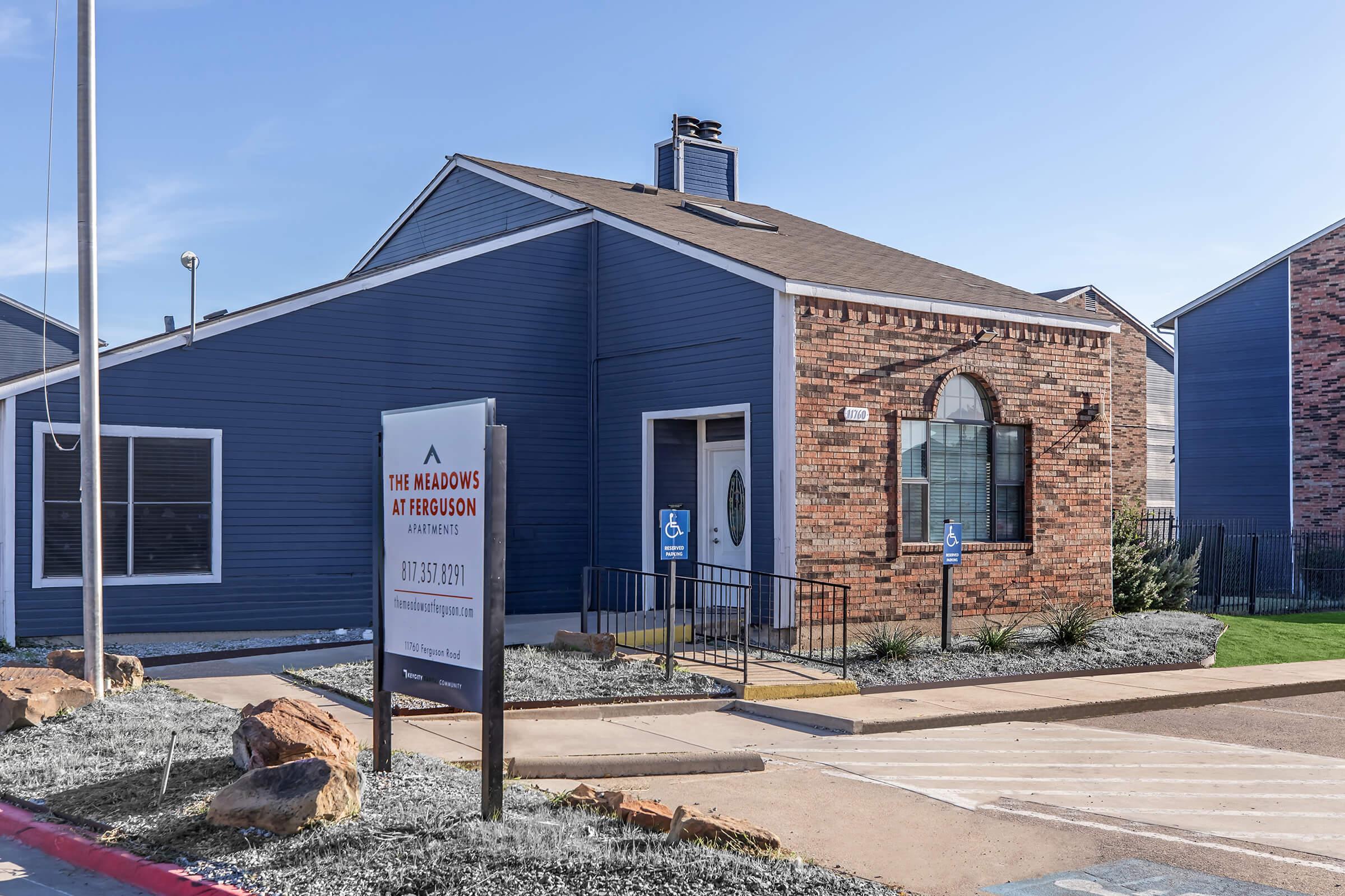 Exterior view of a modern apartment building with blue siding and brick accents. A sign in front reads "The Meadows at Ferguson Apartments" along with contact information. The entrance features a small ramp, and there are landscaped areas with rocks and plants.