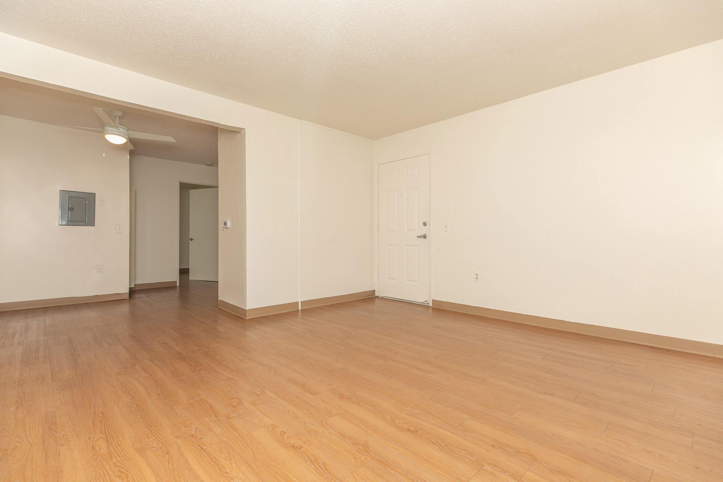Interior view of an empty room with a light-colored wall and hardwood floor. There is a wall-mounted mirror on the left and an entrance door straight ahead. A ceiling fan is visible in the corner, and another room can be seen through an open doorway. The space is bright and uncluttered.