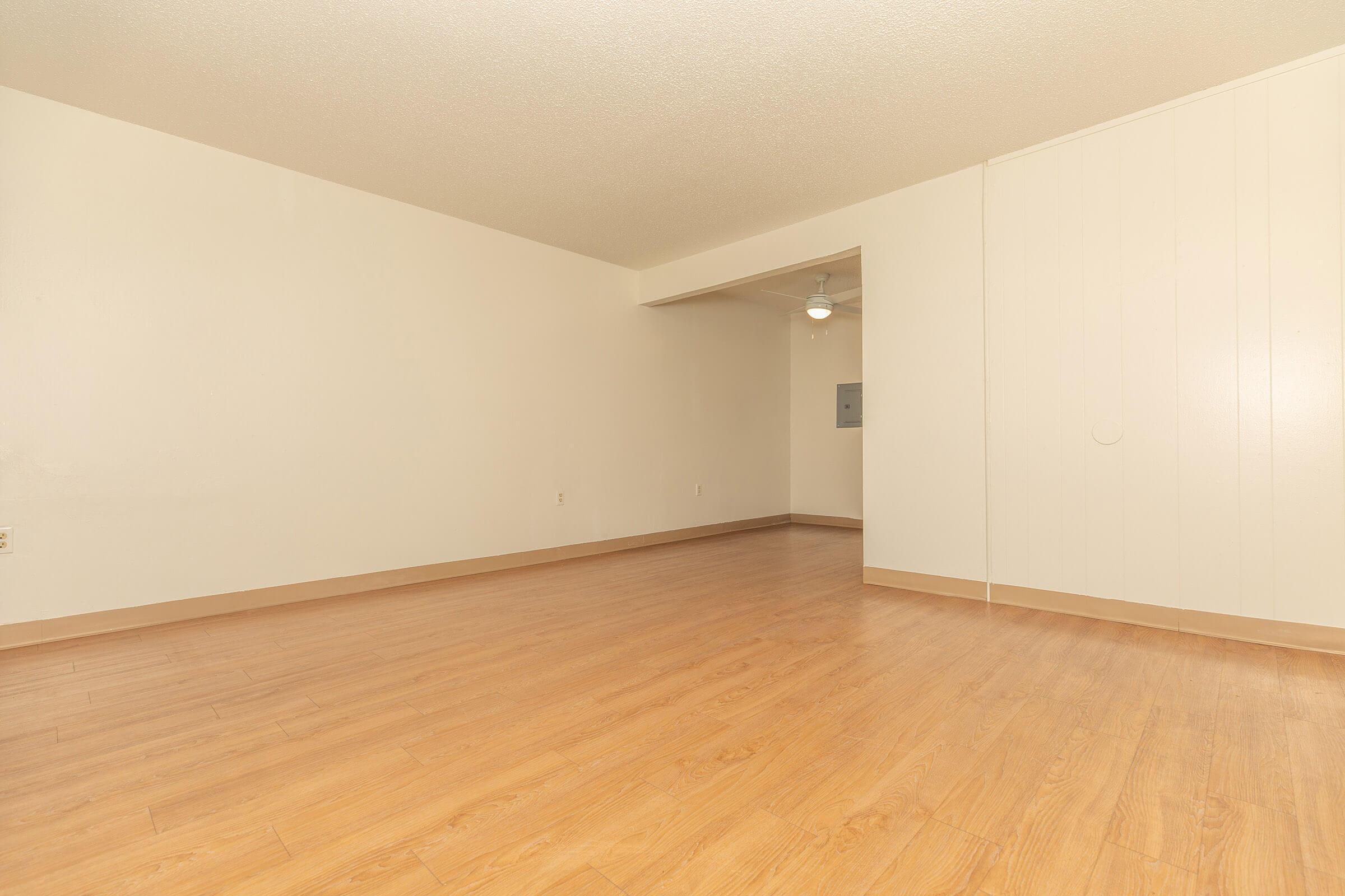 A spacious, empty room with light-colored walls and a wood-like floor. A ceiling fan is visible in the background, and there is a small wall-mounted electrical panel. The atmosphere is bright and inviting, with plenty of natural light illuminating the space.