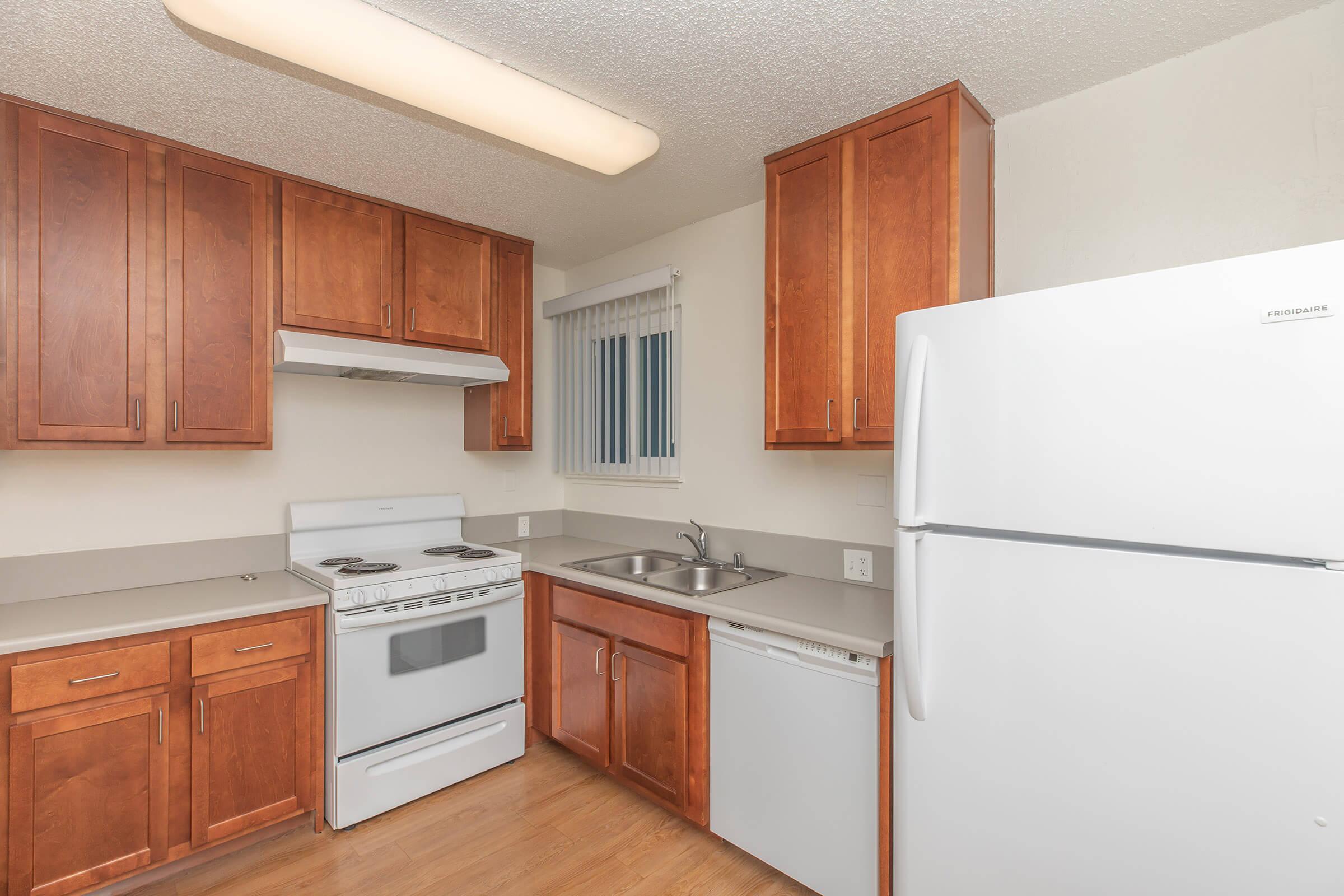 Kitchen featuring wooden cabinets, white appliances including a stove, oven, dishwasher, and refrigerator, a stainless steel sink, and a countertop. The space has laminate flooring and a window with blinds, providing natural light.