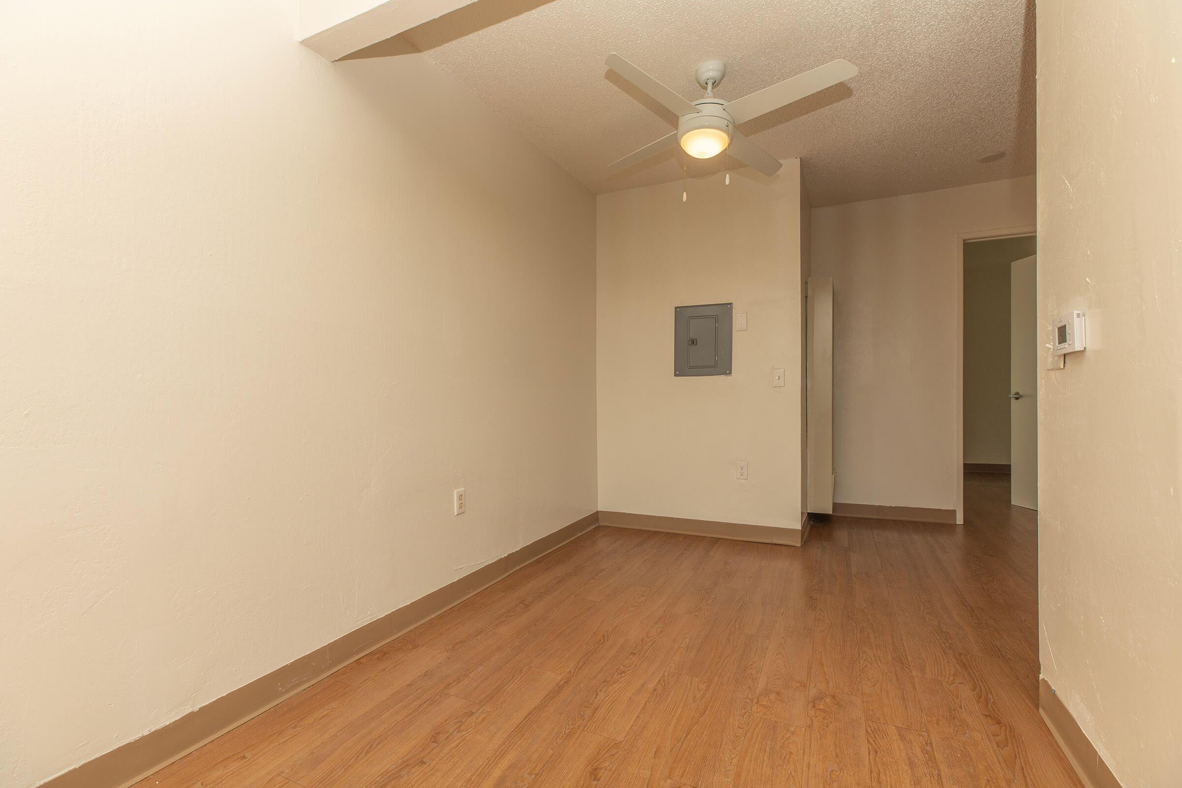 A small room with light-colored walls and hardwood-style flooring. There is a ceiling fan installed, a wall-mounted electrical panel, and a doorway leading to another space. The room is empty with no furniture, providing a clean and minimalistic look.