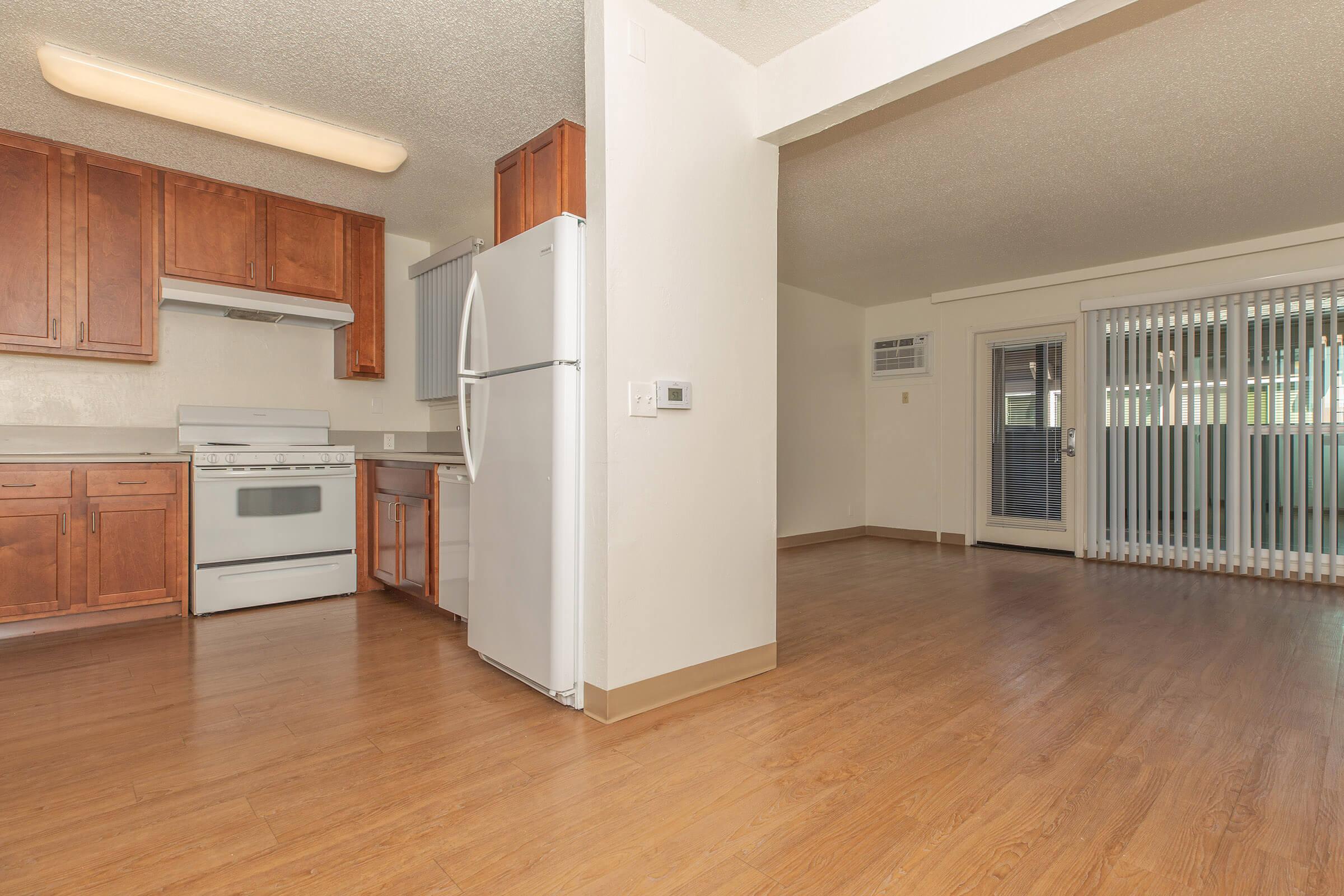 A bright, open kitchen and living area featuring wooden cabinetry, a white refrigerator, and a gas stove. The flooring is a light wood laminate. Towards the back, there is a sliding glass door leading to an outdoor space, with natural light streaming into the room.