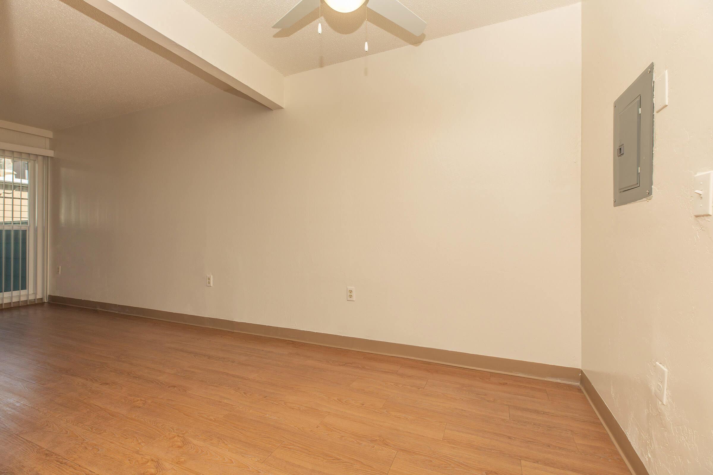 An empty room with light-colored walls and a light wood laminate floor. A ceiling fan is visible above, and there’s an electrical panel on the right wall. The room has large windows allowing natural light, and one side shows a sliding door leading to a balcony or outdoor area.