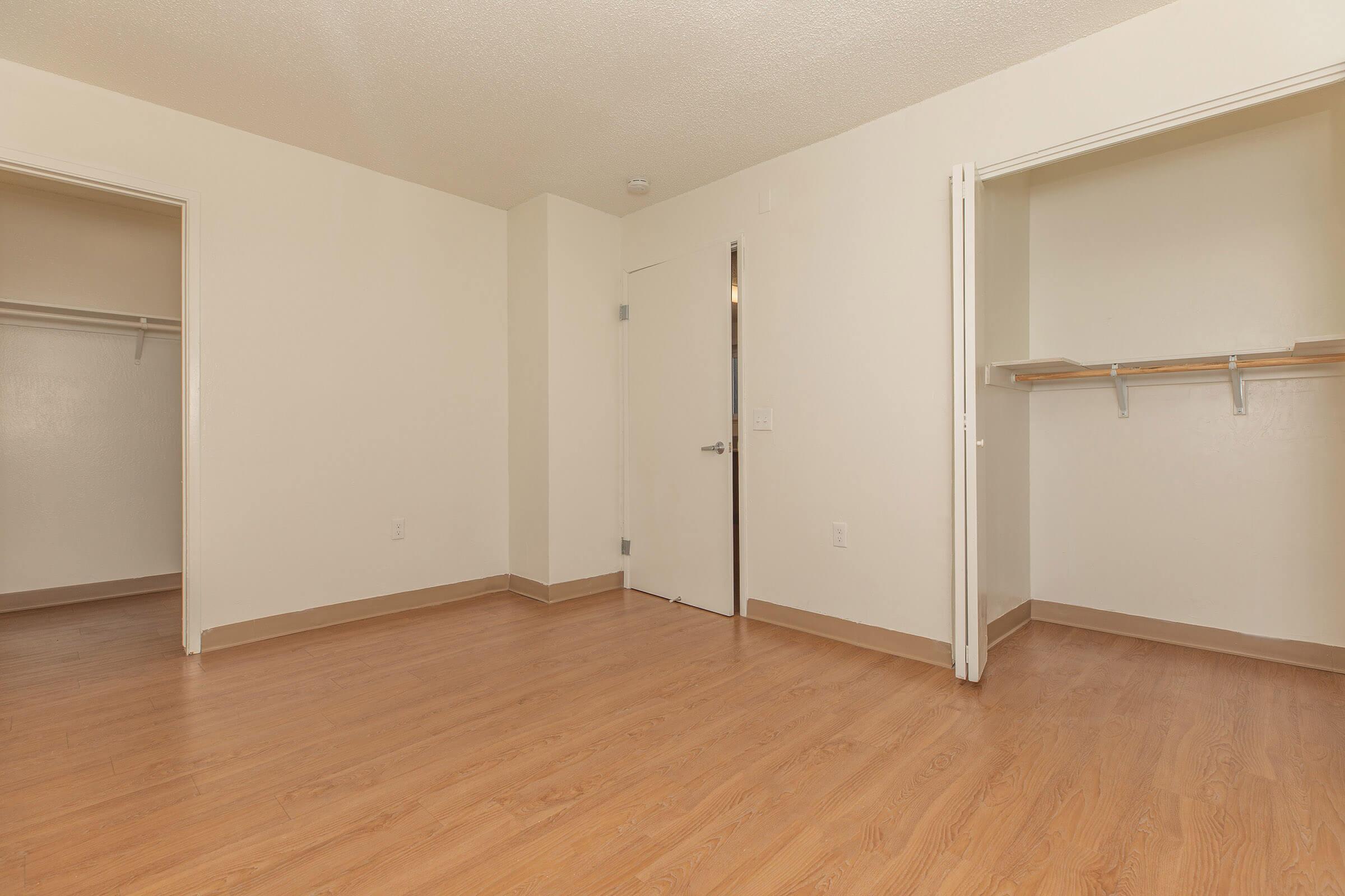 A spacious, empty room with light-colored walls and wooden flooring. Two open closet spaces are visible on the left and right sides of the image. A door is closed at the back, leading to another area. The overall atmosphere is bright and minimalistic, ideal for various design possibilities.
