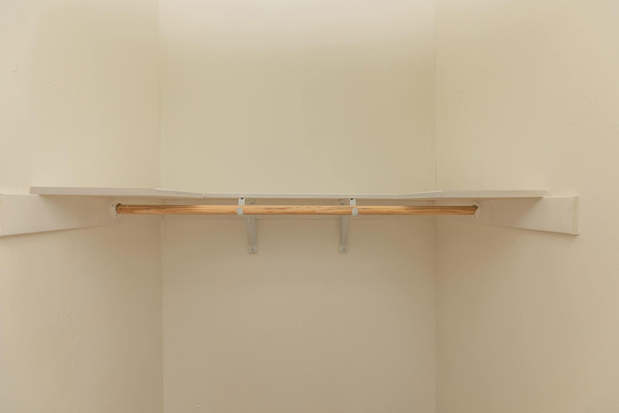 A minimalist closet with a wooden hanging rod and two shelves above it, all set against a plain, light-colored wall. The space is empty, providing a clear view of the hanging area and shelves, emphasizing simplicity and organization.