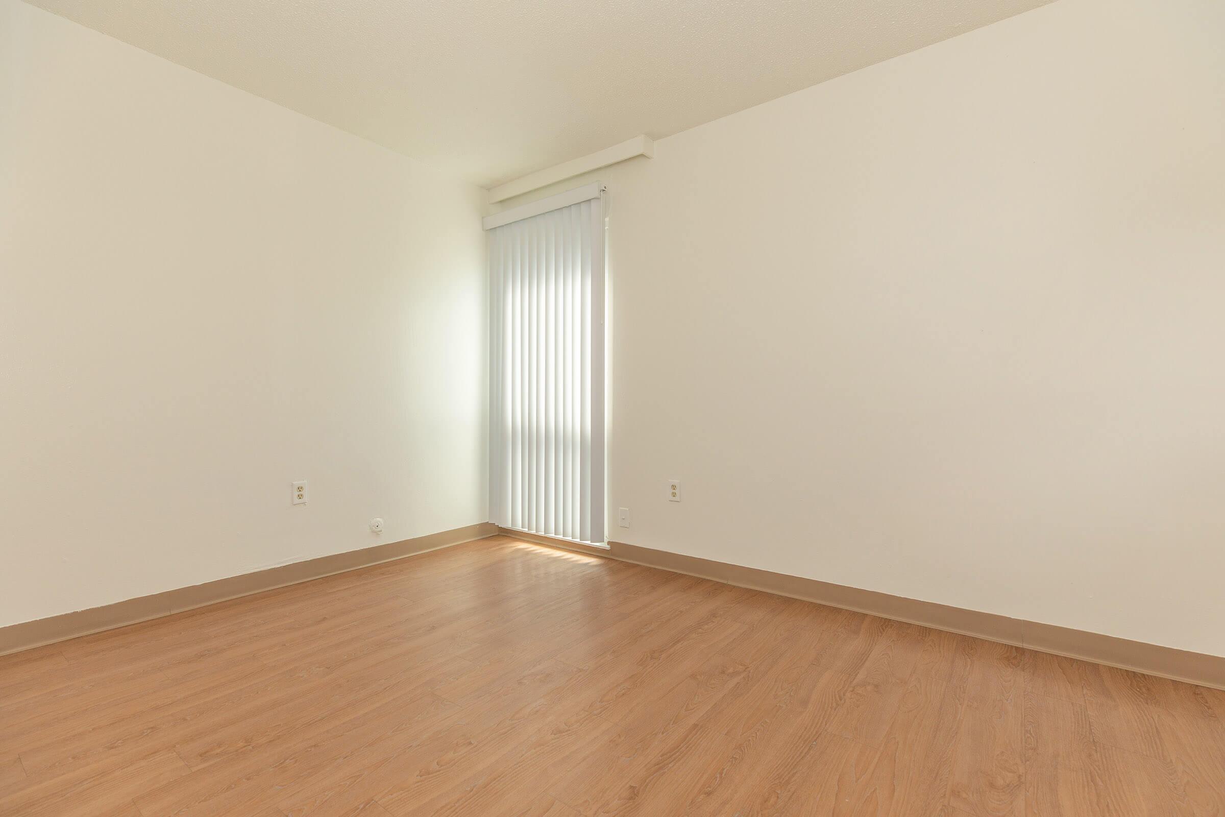An empty room with light-colored walls and a wooden floor. There is a window covered by vertical blinds allowing natural light to enter. The space is minimalist, creating a sense of openness.