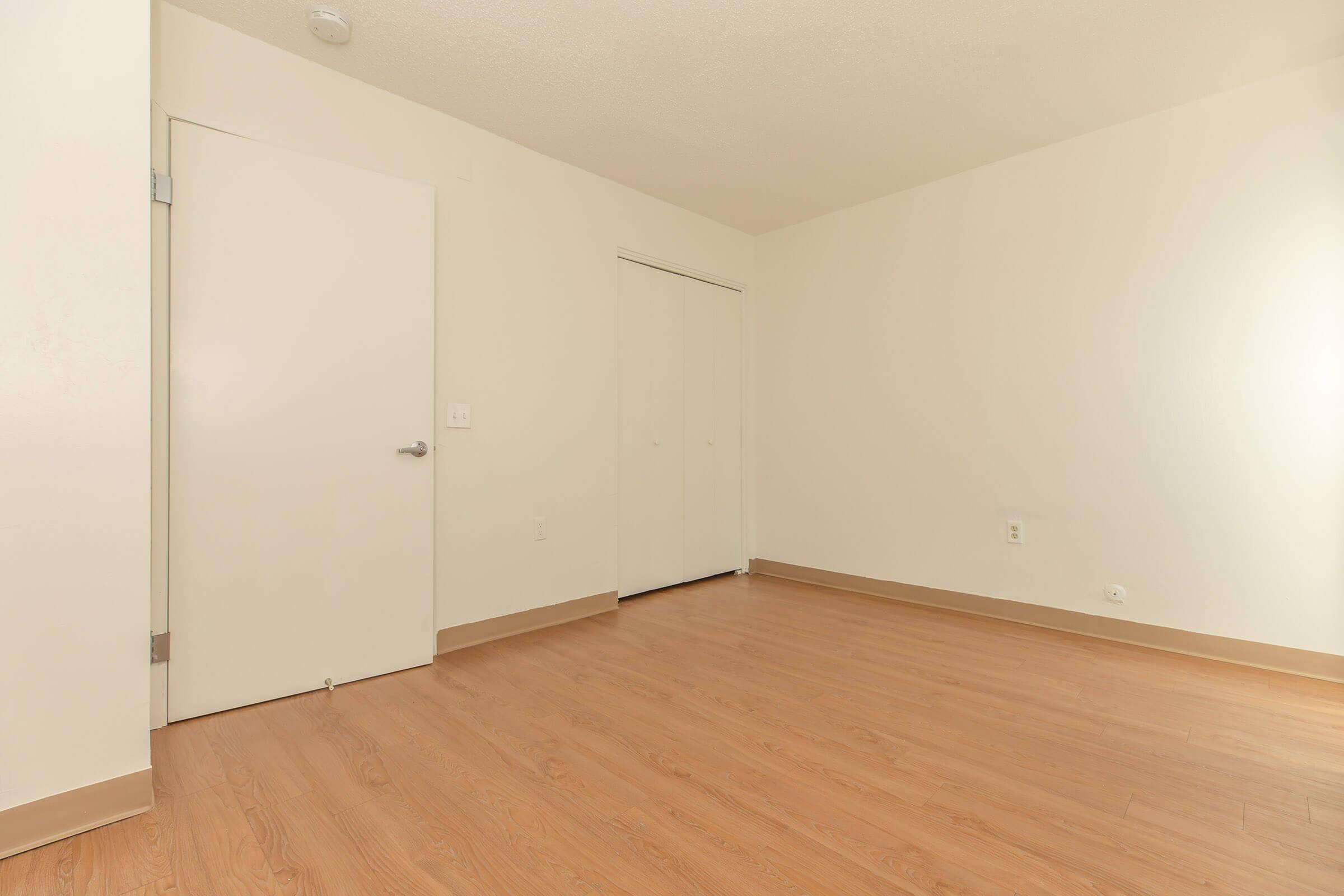 Empty room featuring light-colored walls and wooden flooring. There is a closed white door on the left and a closet with sliding doors on the right. The room is well-lit with natural light, creating a spacious and minimalist atmosphere.
