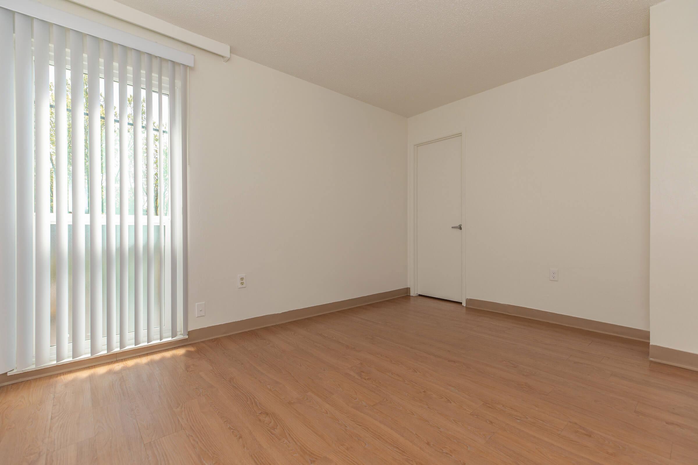 A spacious, empty room with light-colored walls and wooden flooring. A large window with vertical blinds allows natural light to enter, and a closed door leads to another space. The room is unfurnished and includes a neutral color palette, giving it a clean, minimalist look.