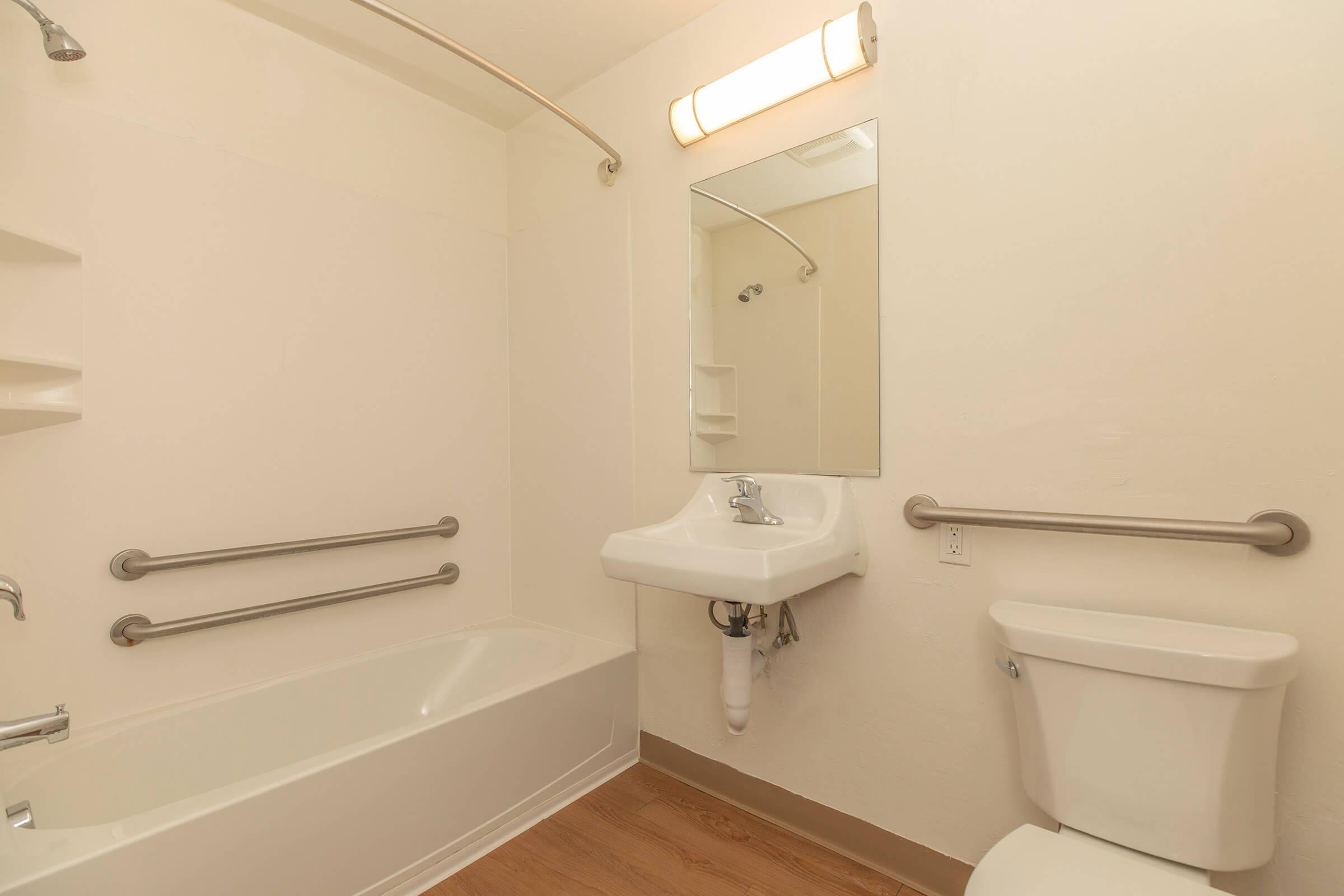 A clean and simple bathroom featuring a bathtub with a shower, a sink with a mirror above, a toilet, and grab bars for safety. The walls are painted light, and the flooring is a light-colored wood. The lighting is provided by a wall-mounted fixture.