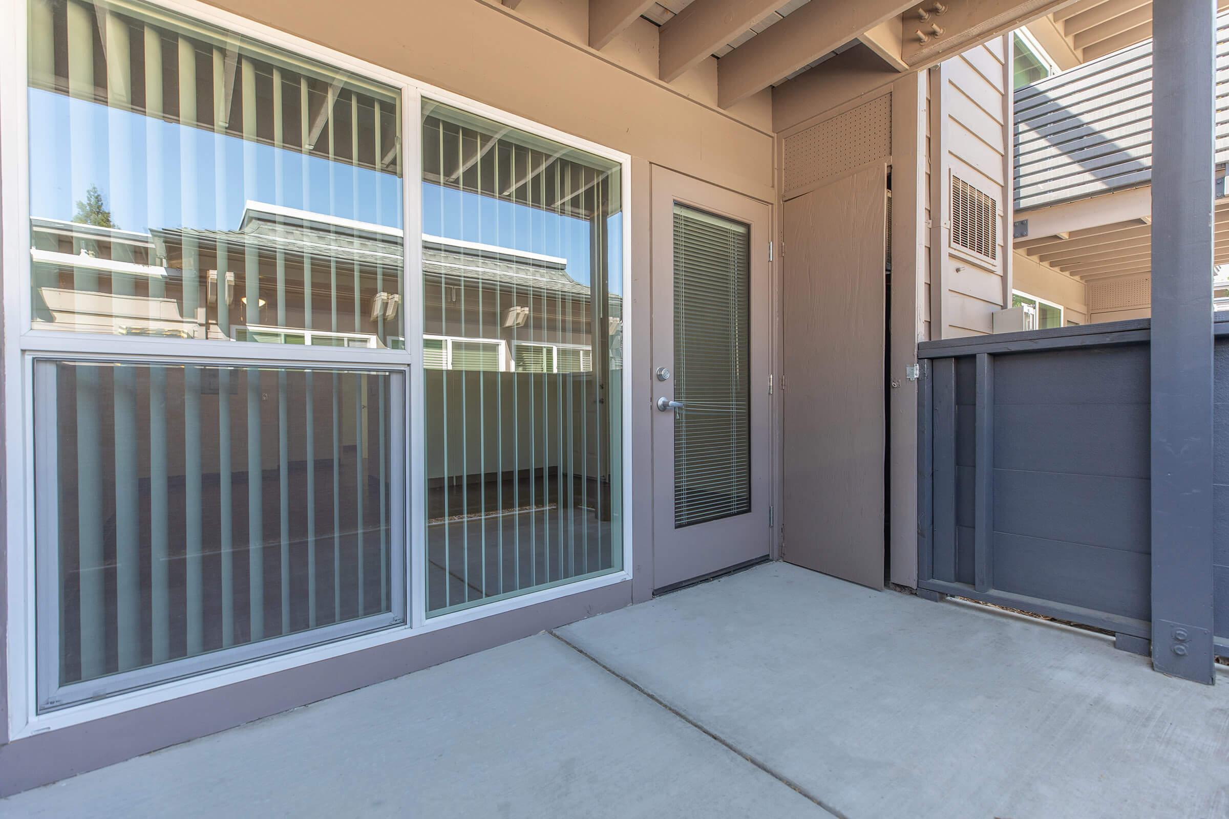 A view of a modern entrance with sliding glass doors and vertical blinds, leading to a patio area. The exterior features a clean, concrete floor and nearby wooden fencing. Natural light reflects off the glass, giving the space a bright, inviting appearance.